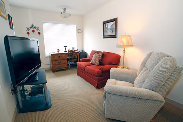 A small living room with a red loveseat, a beige recliner, a wooden desk with a chair near a window with blinds, a floor lamp, and a flat-screen TV on a stand.