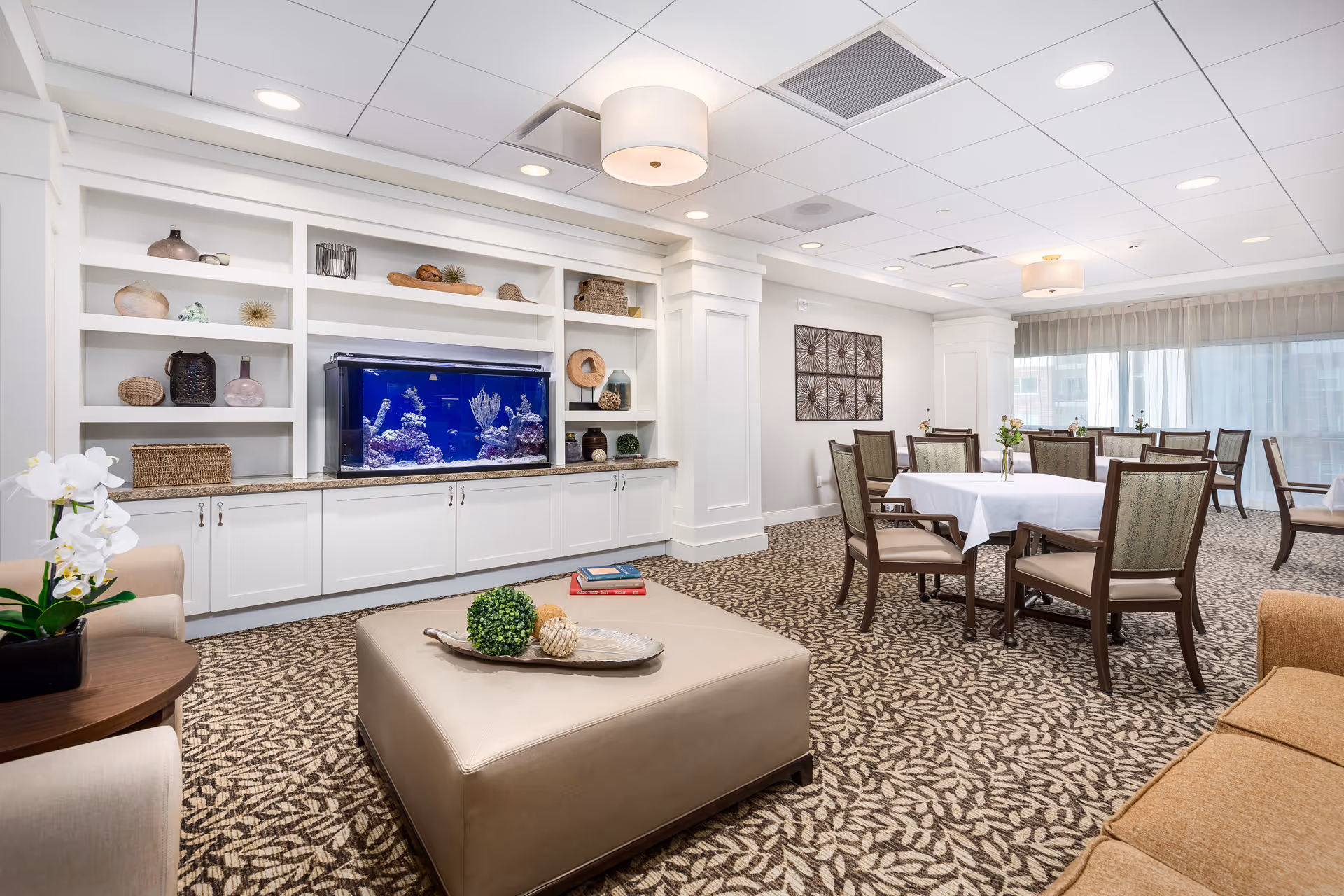 A bright and spacious common area in a senior living facility featuring a large built-in white shelving unit with decorative items and a large aquarium. The room has a patterned carpet, several tables with chairs, and comfortable seating including a beige ottoman and a sofa. Large windows with sheer curtains allow natural light to fill the space.
