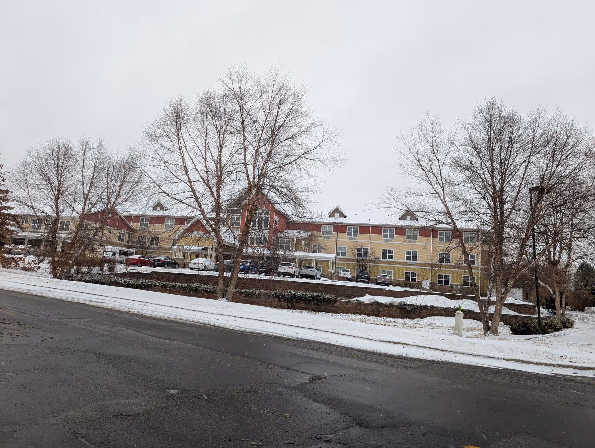 Exterior front view of a multi-story senior living building in winter with snow on the ground, parked cars, and leafless trees.