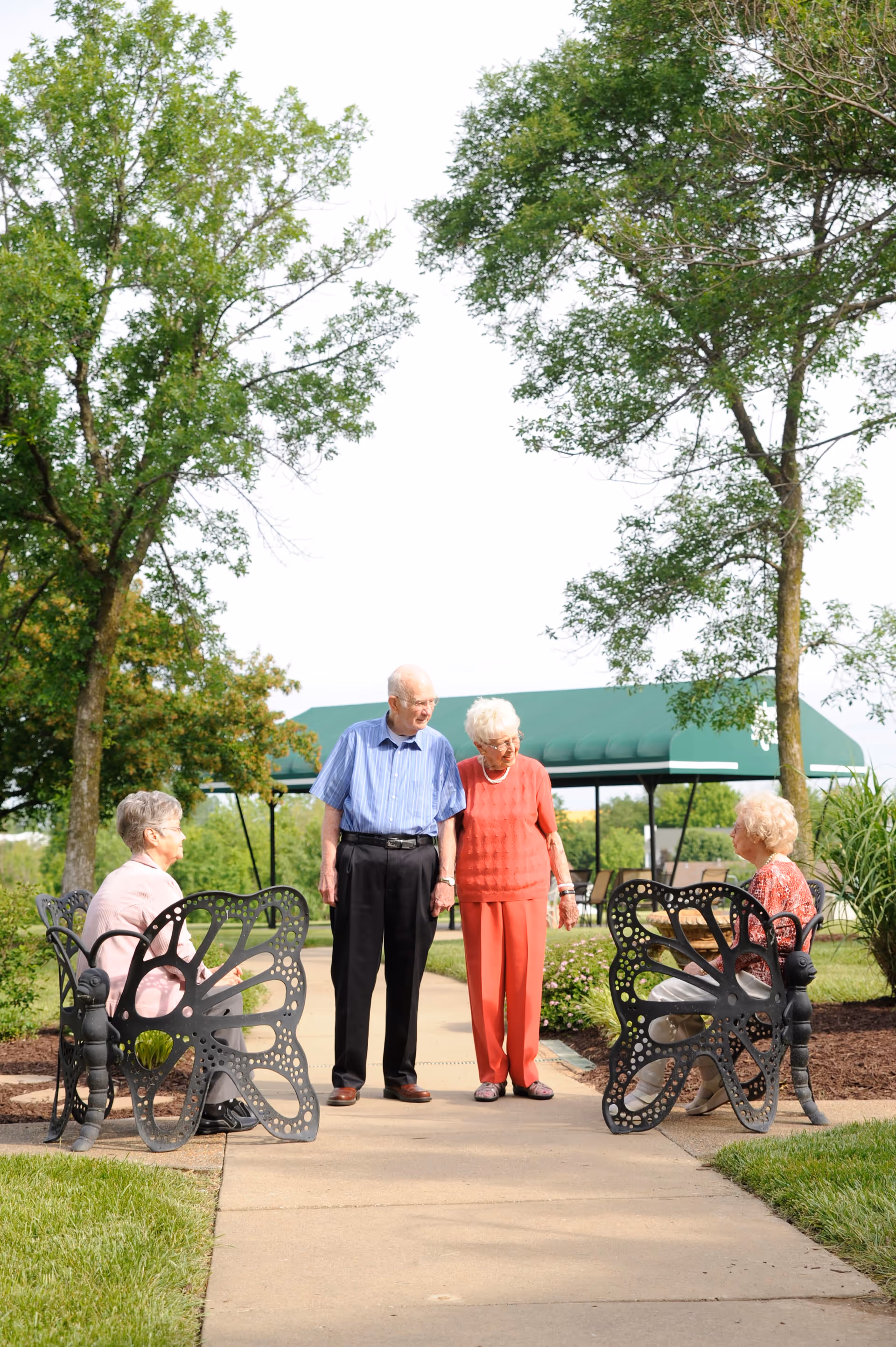 Two elderly women sitting on butterfly-shaped metal benches facing each other on a paved pathway in a garden area, with an elderly man and woman standing and holding hands between them. There are trees and a green canopy structure in the background.