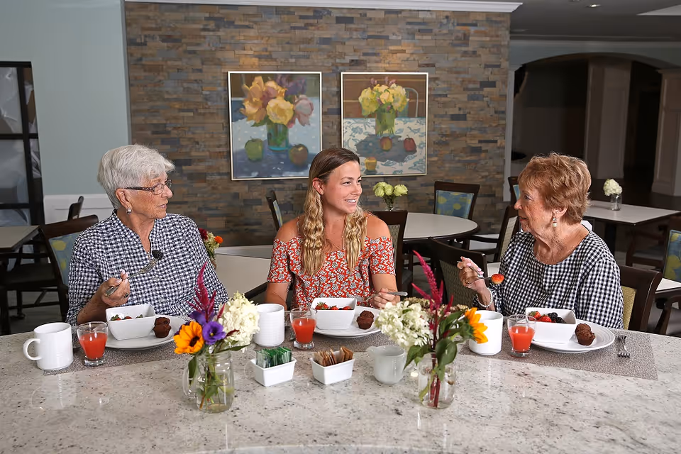 Three women sitting at a dining table in a senior living facility, enjoying a meal together. The table has white mugs, glasses of red juice, bowls of fruit, and small flower arrangements. The background features a stone accent wall with two paintings of flowers and fruit.