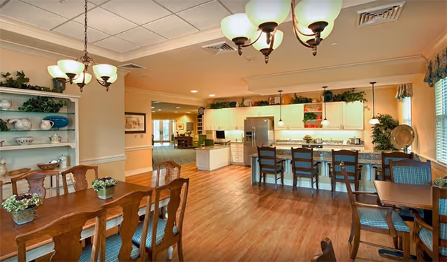 A spacious dining and kitchen area in a senior living facility with wooden floors, multiple dining tables and chairs, a kitchen island with bar stools, white cabinets, and decorative plants and dishes on shelves.