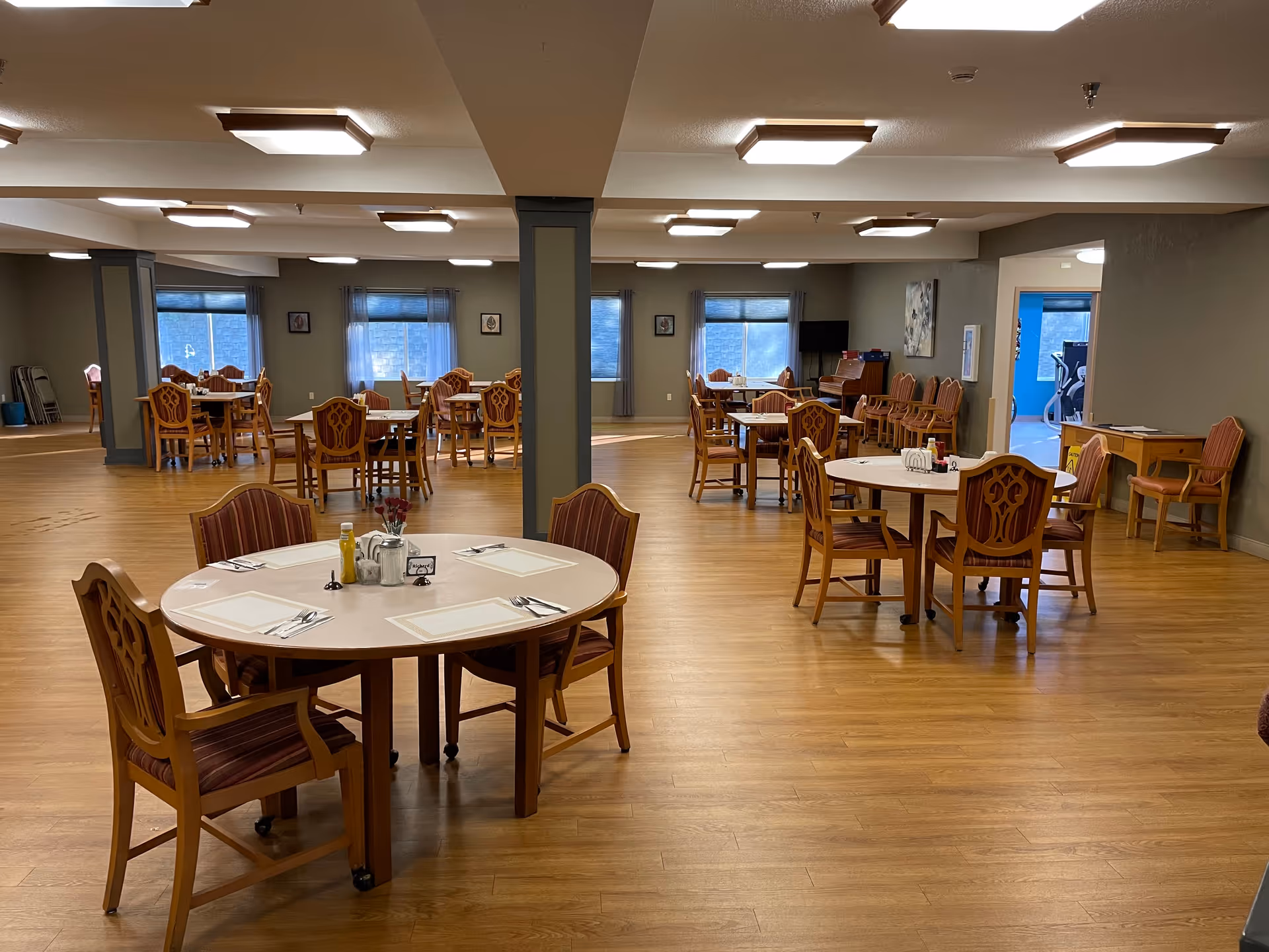 A spacious dining room in an assisted living facility with multiple round tables and wooden chairs arranged neatly. The room has large windows with curtains, wooden flooring, and soft overhead lighting. There is a small desk with chairs in the corner and a glimpse of an adjacent room with exercise equipment.