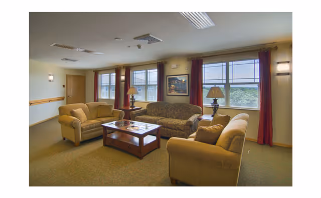 A senior living facility common area with three upholstered sofas arranged around a wooden coffee table. The room has large windows with red curtains, two table lamps on side tables, and a framed painting on the wall. The carpet is greenish, and the walls are light-colored.