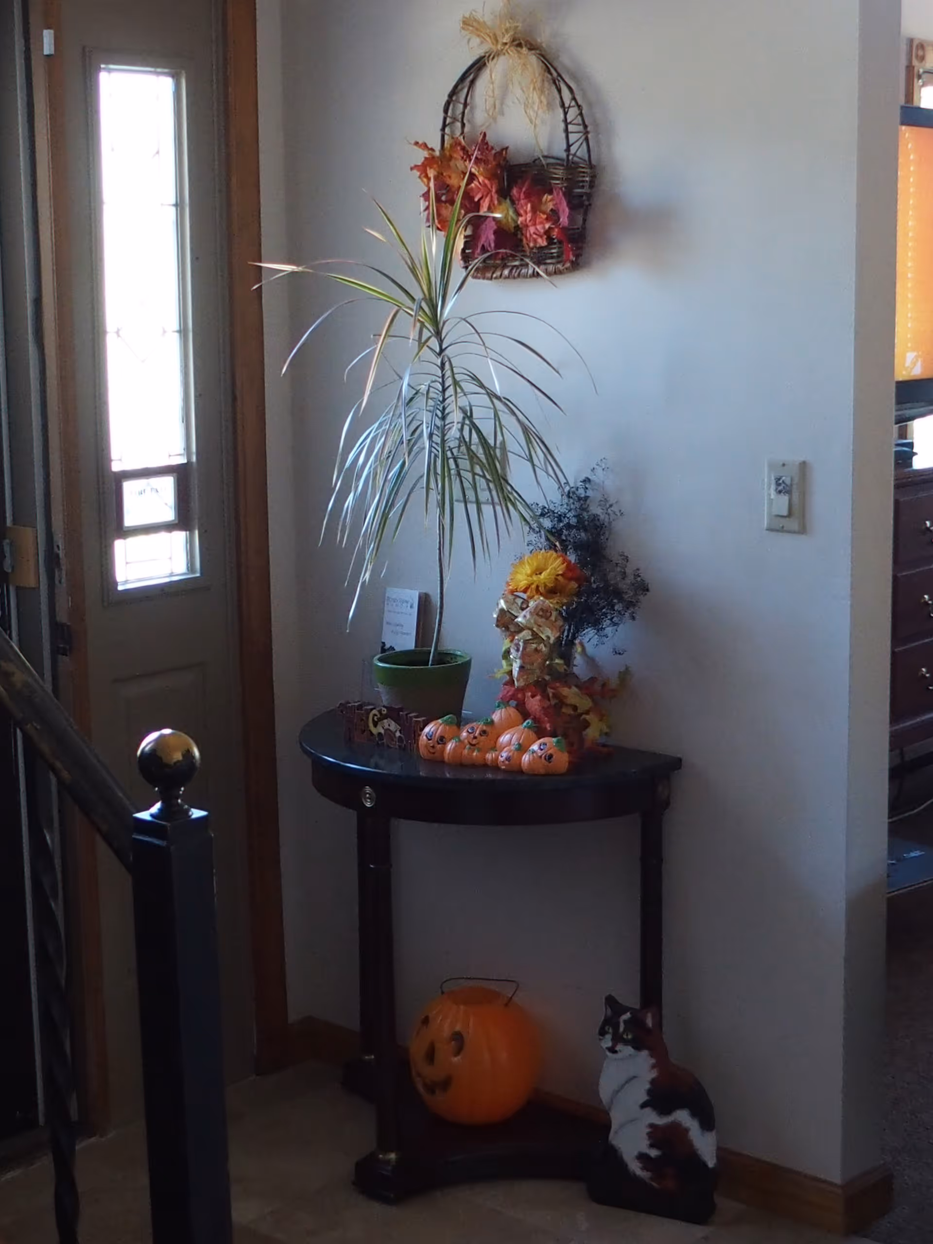 A small dark wooden half-moon table against a white wall near a door with a window. On the table are a potted plant, a small decorative scarecrow, and several small pumpkin decorations. Below the table is a larger pumpkin decoration and a cat figurine. A basket with autumn leaves hangs on the wall above the table.