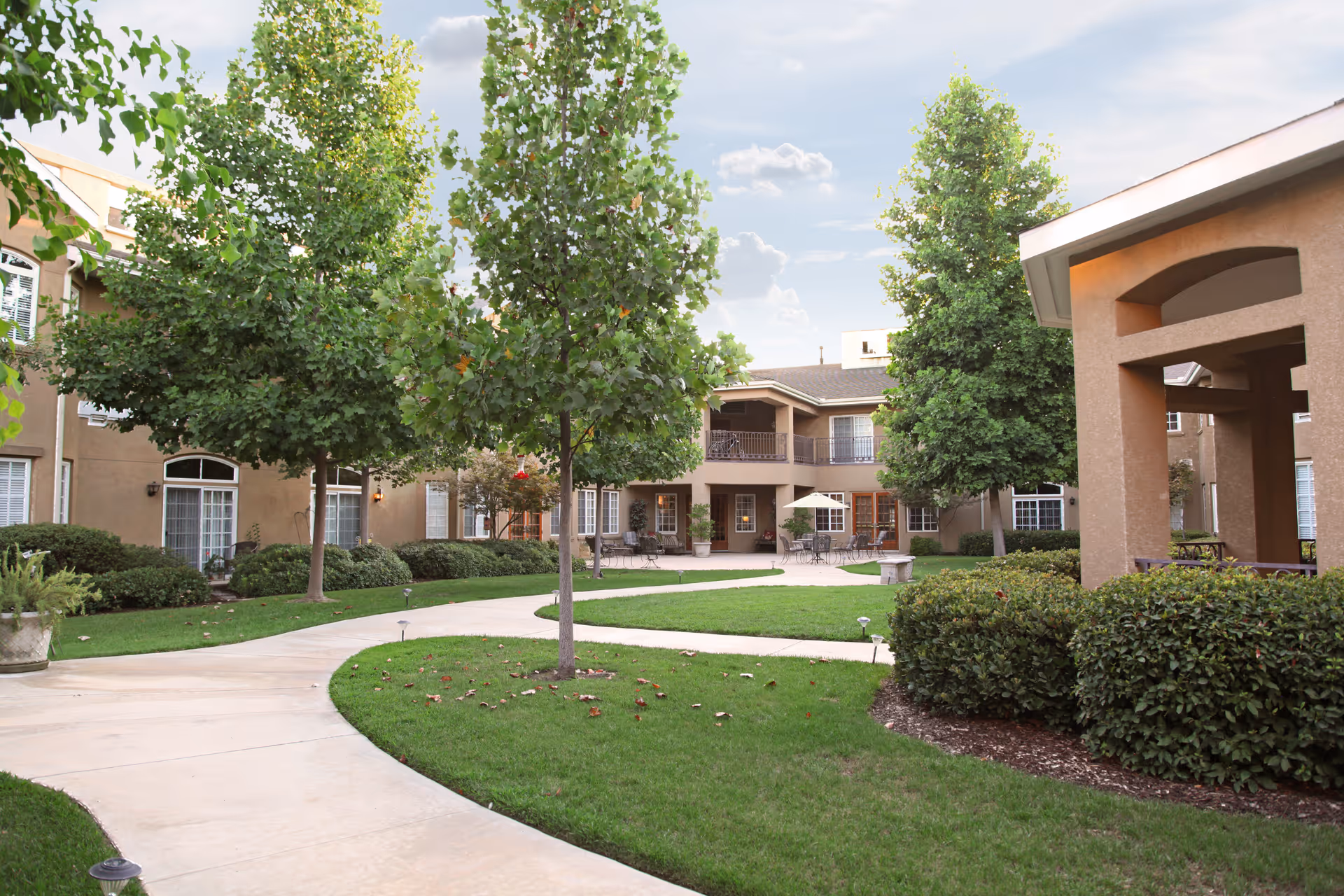 Outdoor courtyard area of a senior living facility with a curved concrete walkway, green grass, several trees, bushes, and a two-story building with balconies and windows surrounding the courtyard. There are patio tables and chairs with umbrellas near the building.