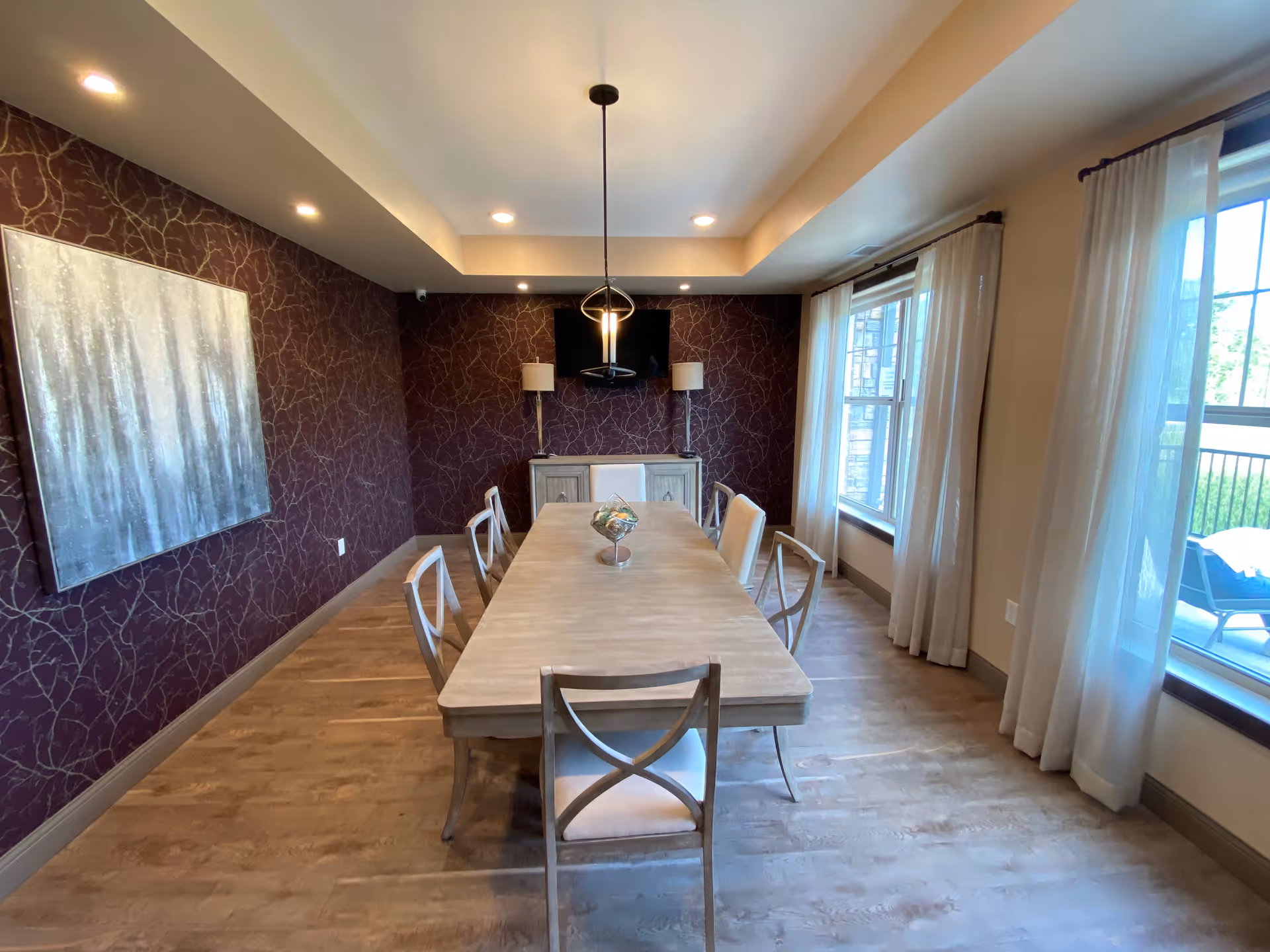 Bright dining room with a long wooden table surrounded by chairs, patterned accent wall, large windows with curtains, and a central pendant light.