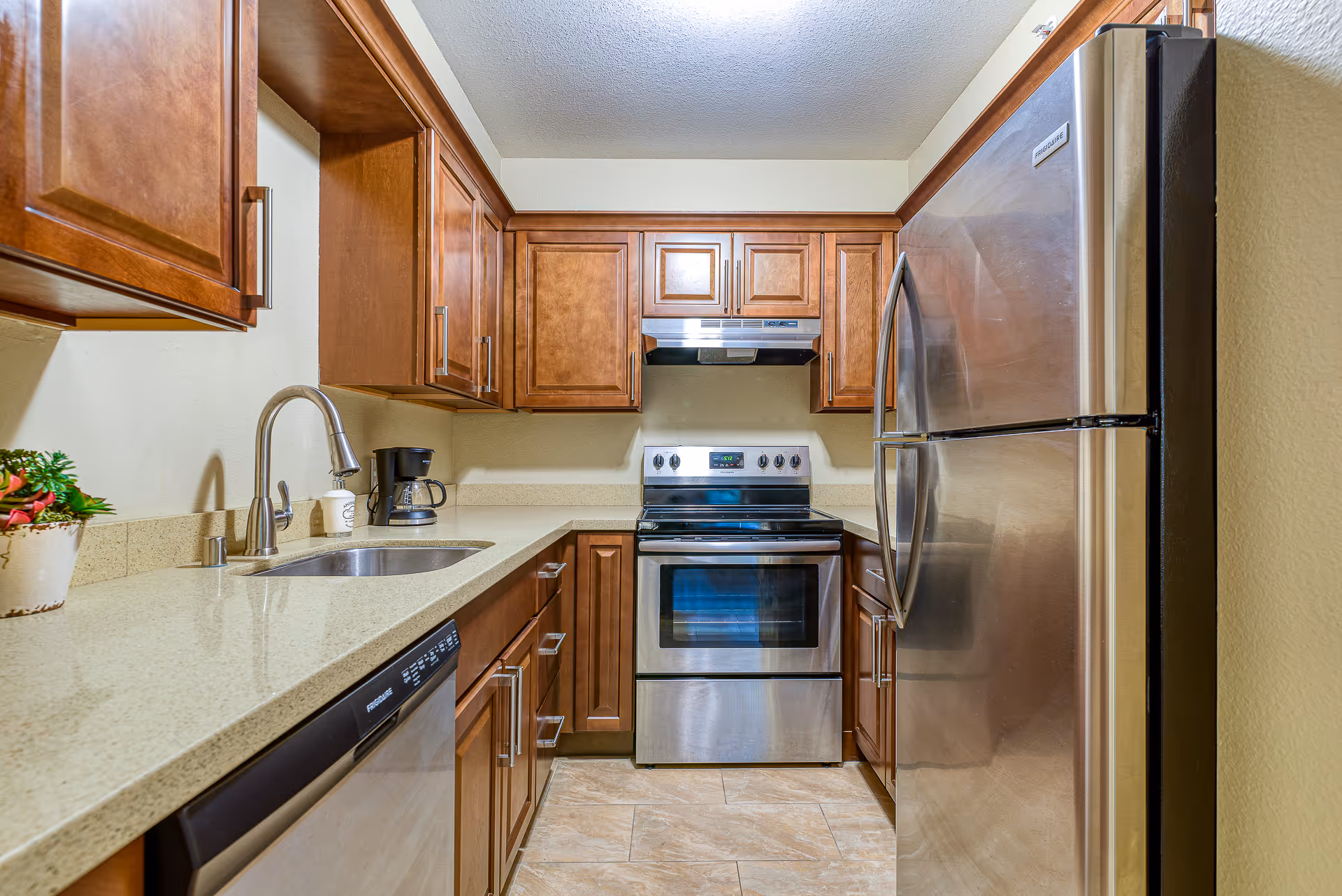 A modern kitchen with wooden cabinets, a stainless steel refrigerator, oven, and dishwasher. The kitchen has a beige countertop with a sink, a coffee maker, and a small potted plant. The floor is tiled and the walls are light-colored.