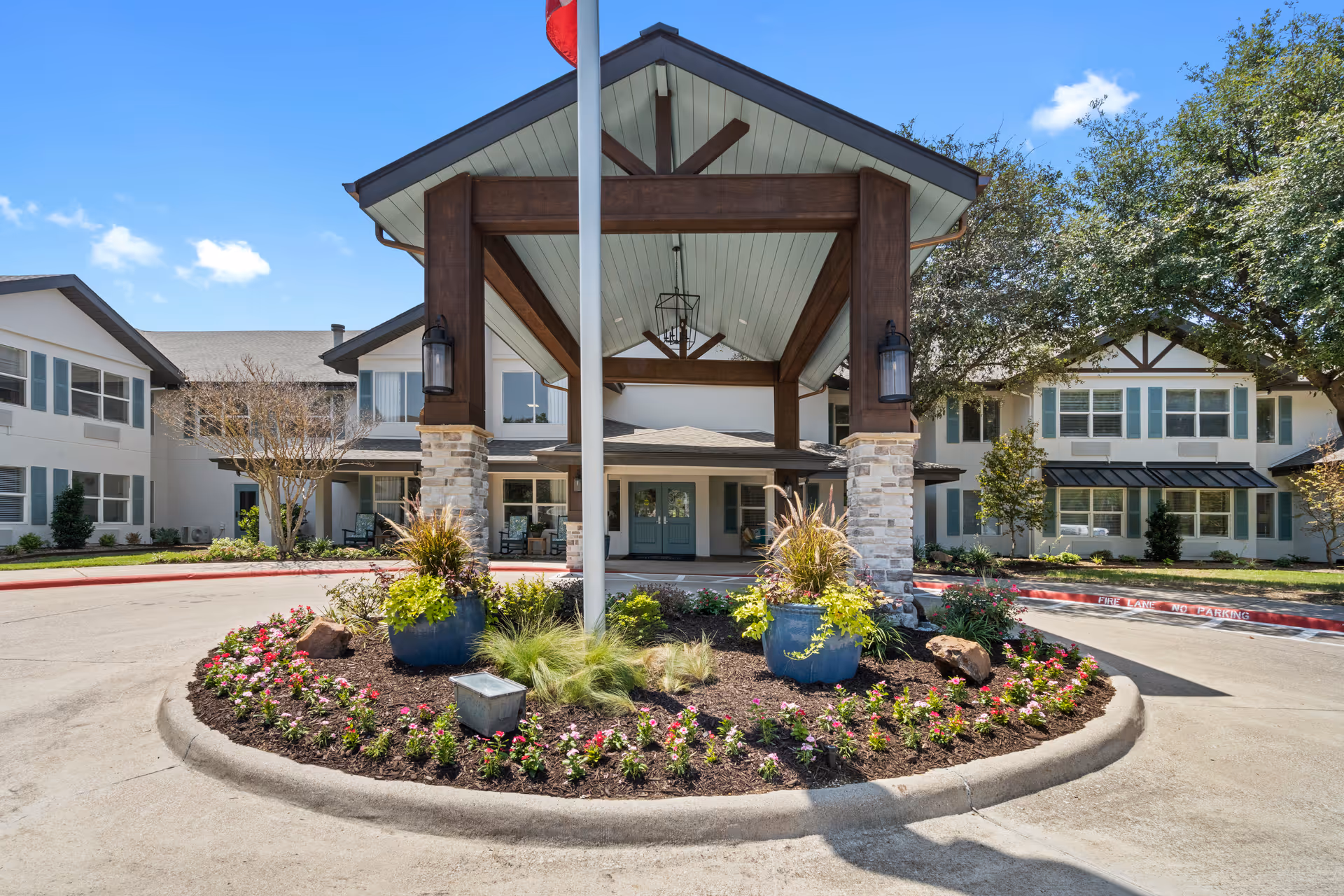 Front entrance of The Grove on Forest Lane senior living facility with a covered driveway, landscaped flower beds, and a flagpole under a clear blue sky.