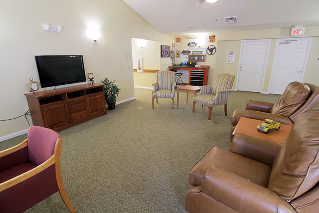 A living room area in a senior living facility with a flat-screen TV on a wooden stand, two striped armchairs around a small wooden table, a brown leather recliner sofa with a toy car on the armrest, and a purple cushioned chair. The room has beige walls, carpeted floor, and various decorations on the far wall including signs and a steering wheel.