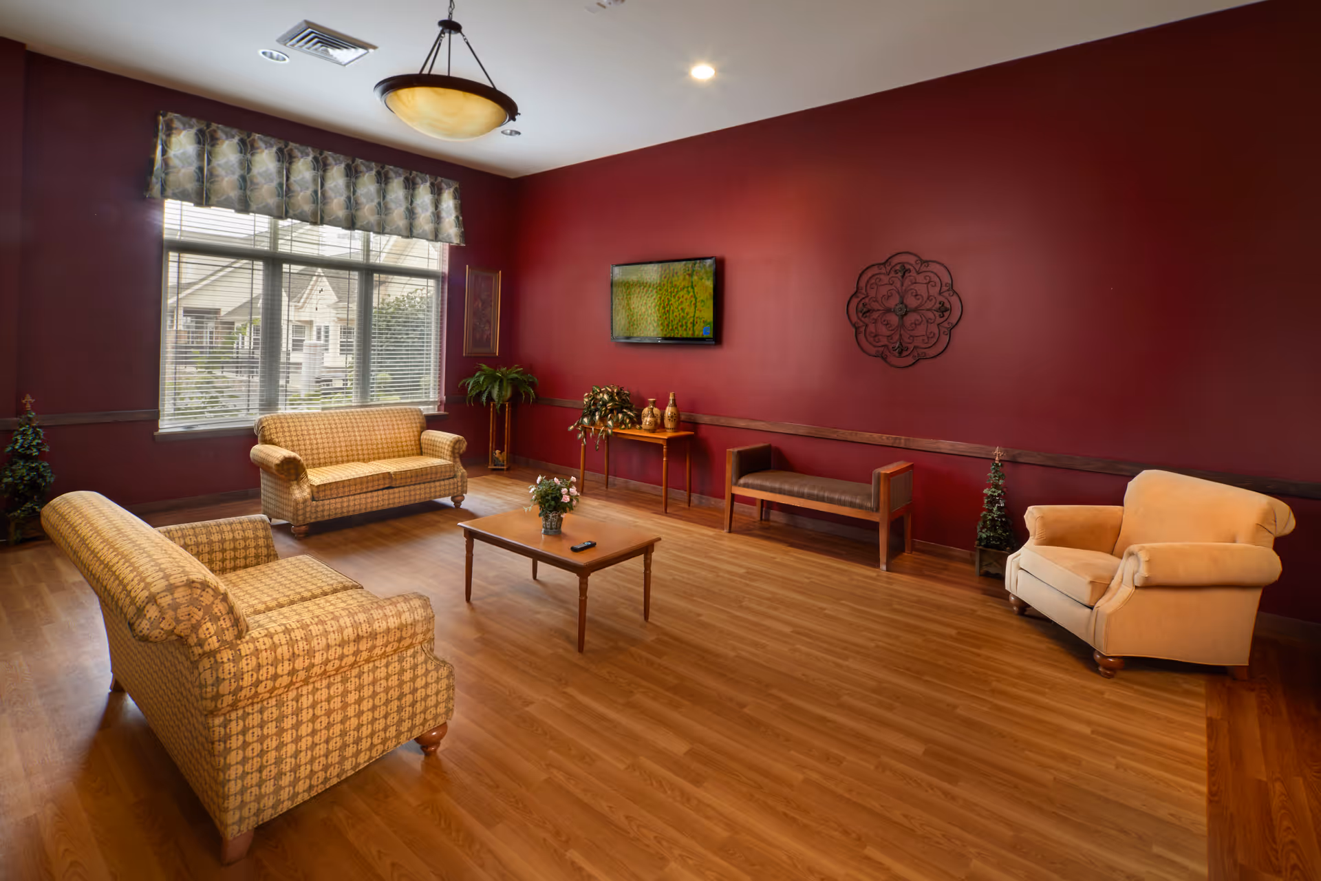 A cozy living room with wooden flooring and deep red walls. The room features two patterned sofas, a beige armchair, a wooden coffee table with a small plant and remote control on it, and a wooden bench against the wall. A flat-screen TV is mounted on the wall above a small table with decorative items. Large windows with patterned valances allow natural light to brighten the space, and there are several potted plants placed around the room.