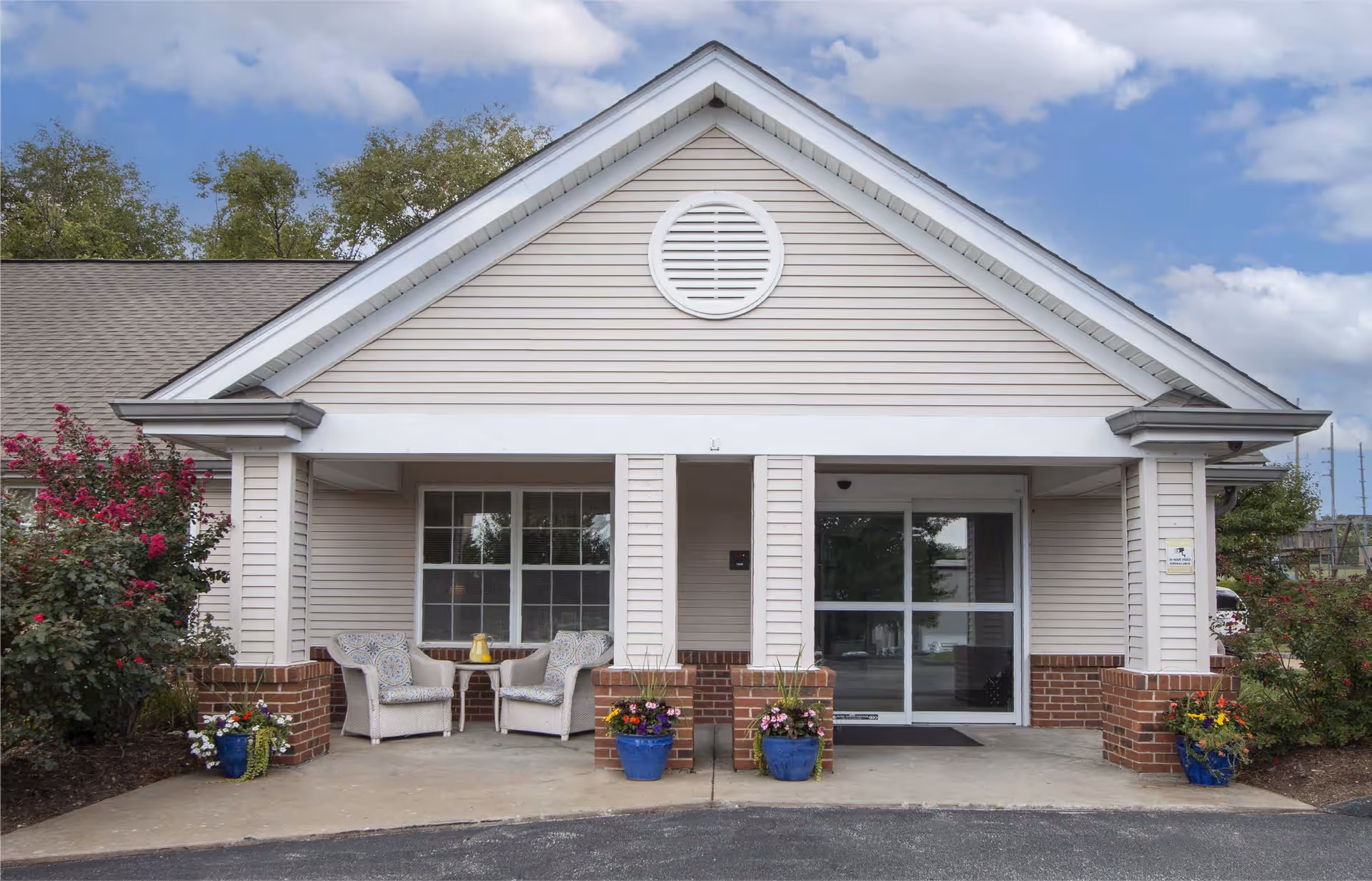Front entrance of a single-story building with beige siding and a gabled roof. There are two white wicker chairs with patterned cushions and a small table with a yellow pitcher on the left side of the entrance. Four blue flower pots with colorful flowers are placed near the brick pillars. The entrance has a glass sliding door and a window beside it. The sky is partly cloudy.