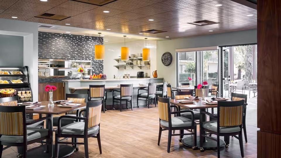 Well-lit dining area with tables and chairs, place settings and vases of flowers, and a serving counter with an open kitchen in the background.