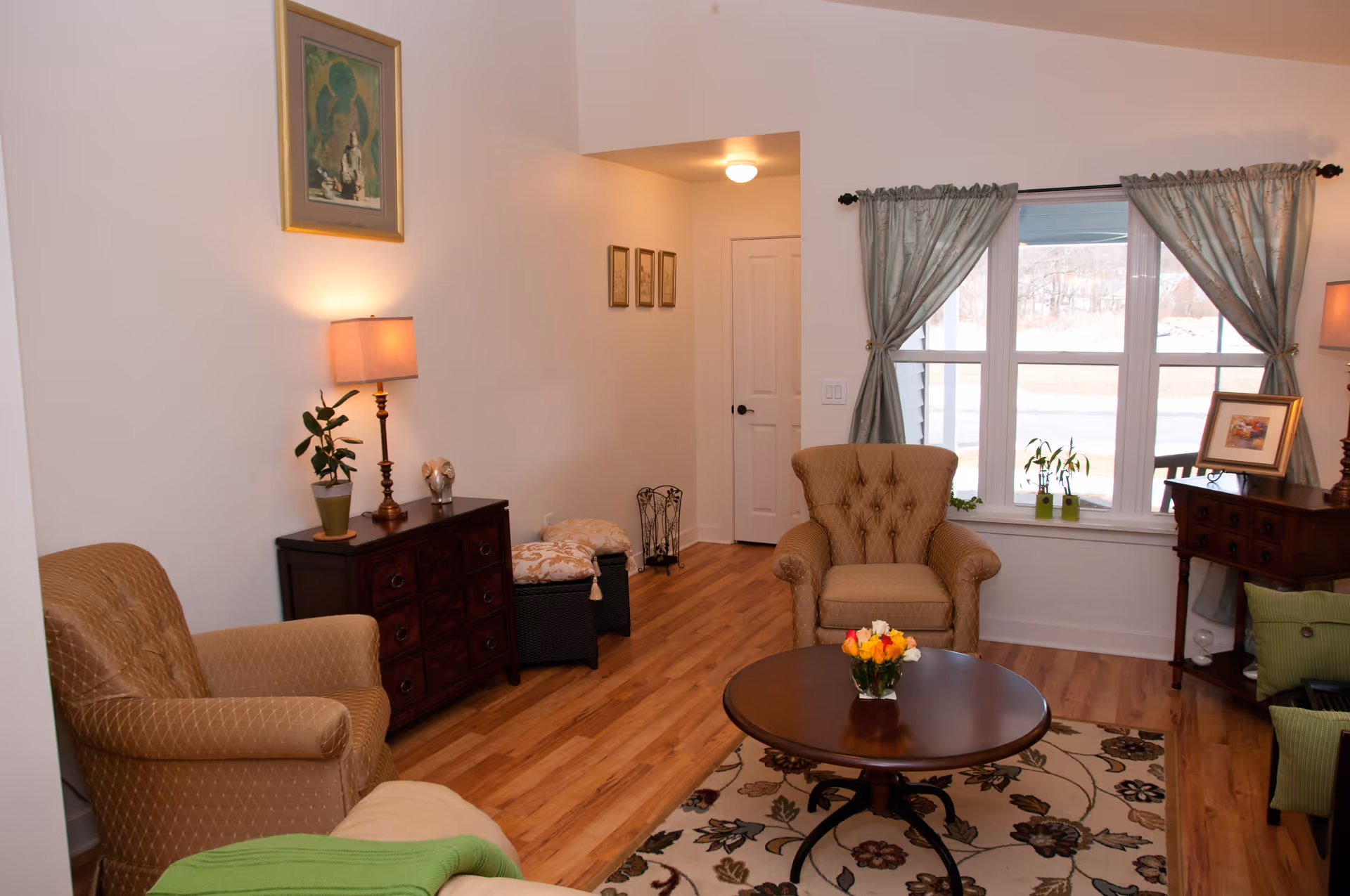 A cozy living room with two upholstered armchairs, a round wooden coffee table with a small flower arrangement, a patterned area rug, wooden flooring, two side tables with lamps and plants, a window with light green curtains, and framed artwork on the walls.