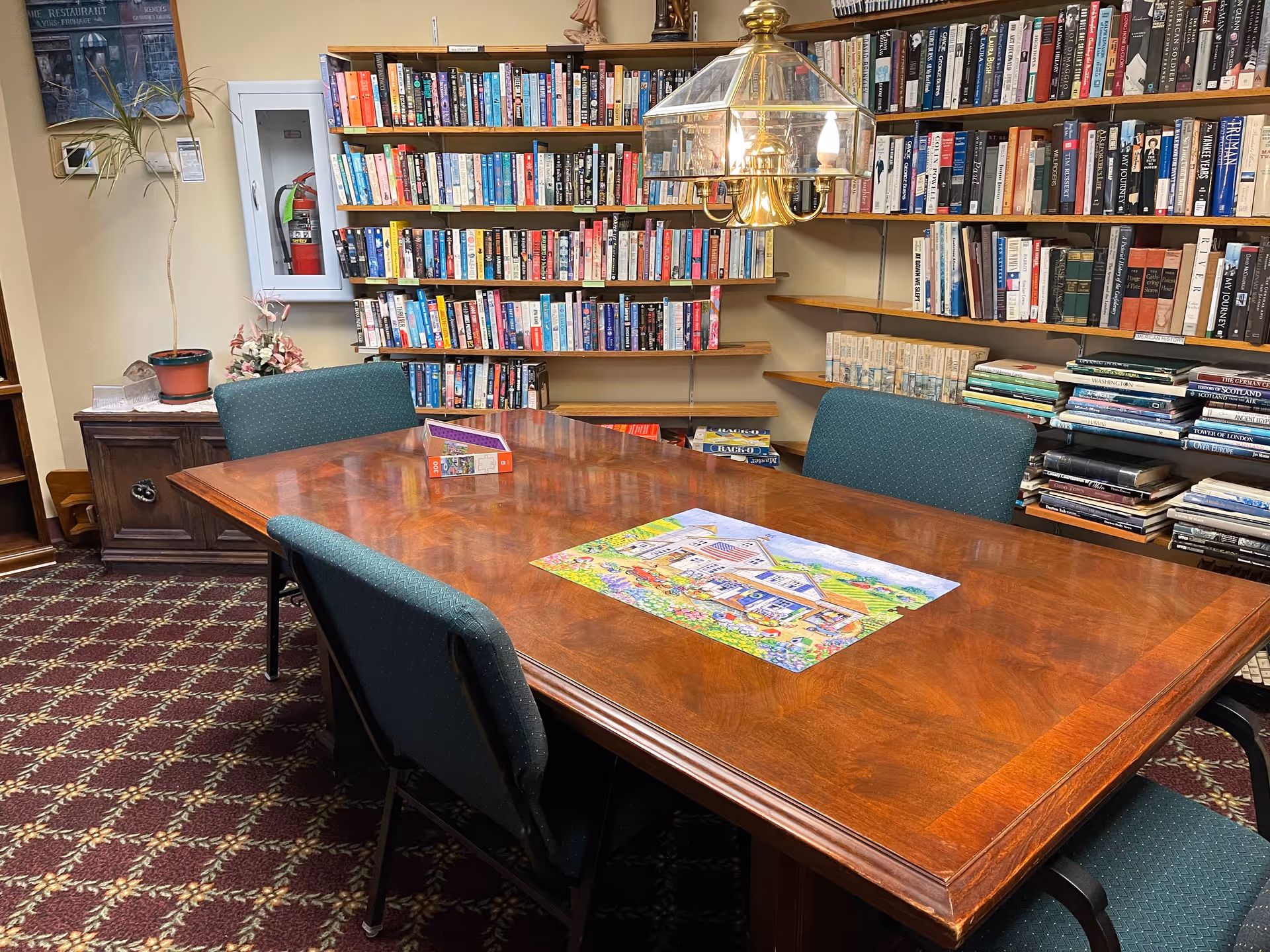 A cozy library room with a large wooden table in the center surrounded by green upholstered chairs. The table has a colorful puzzle and a box on it. Behind the table, there are shelves filled with books. A brass chandelier with glass shades hangs above the table. The room has patterned carpet and a small cabinet with a potted plant and flowers on it.