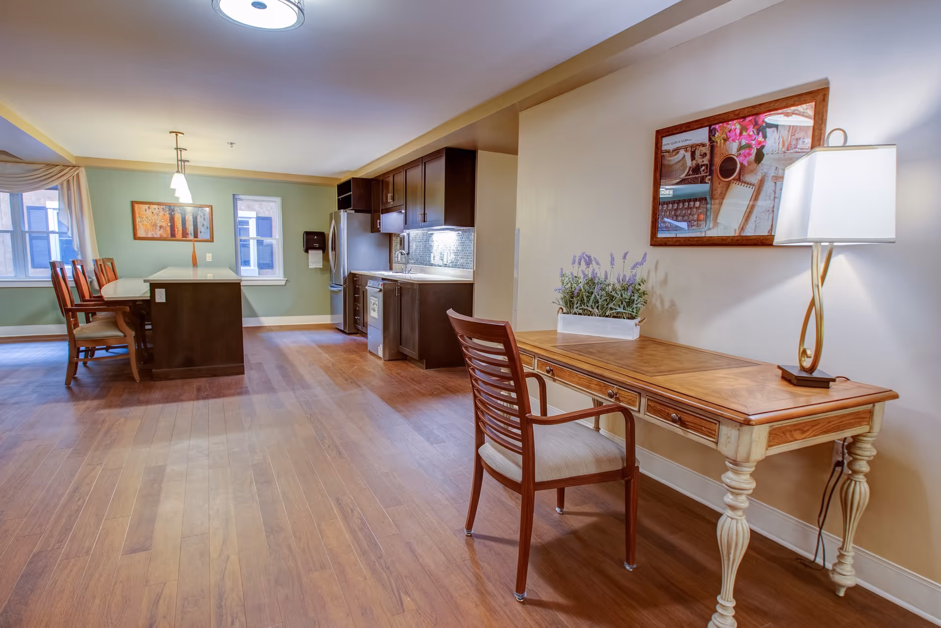 A spacious interior room featuring a kitchen area with dark wood cabinets, stainless steel refrigerator, and dishwasher. Adjacent to the kitchen is a dining area with a table and chairs near two windows. In the foreground, there is a wooden desk with a chair, a table lamp, and a decorative plant. The walls are painted in light colors with framed artwork hanging.