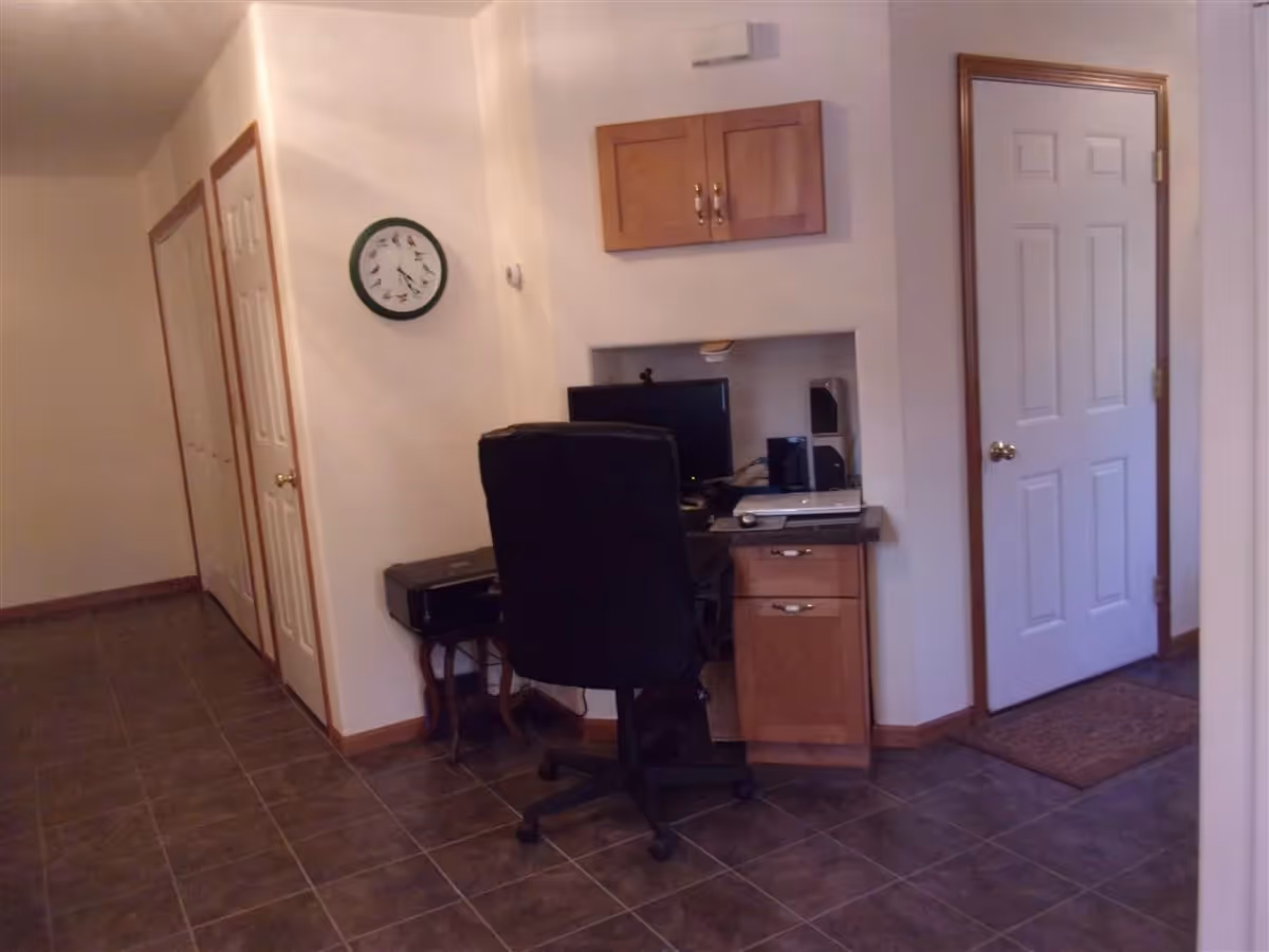 A small home office area with a black office chair in front of a desk that has a computer monitor, laptop, and speakers. Above the desk is a wooden cabinet mounted on the wall. To the left, there is a wall clock with bird illustrations and two closed white doors with wooden trim. The floor is tiled with dark tiles, and there is a white door with a brass knob on the right side of the image.