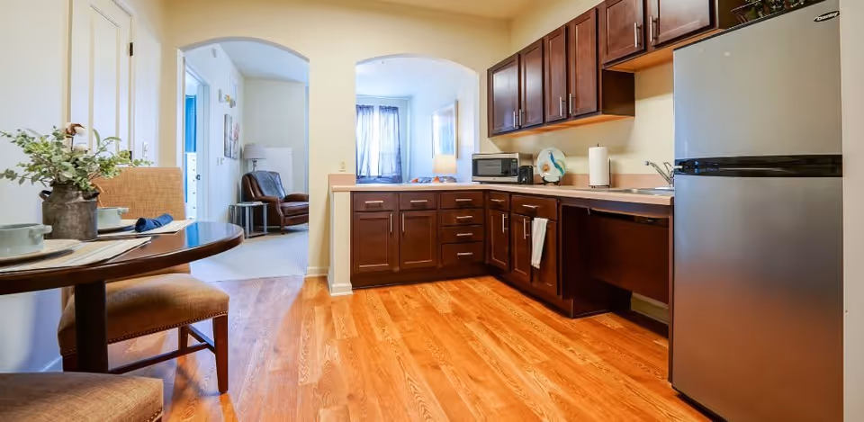 A kitchen area with wooden cabinets, a stainless steel refrigerator, a microwave, and a small round dining table with two chairs. The floor is wooden, and there are two arched doorways leading to a living area with a leather chair and a window with curtains.