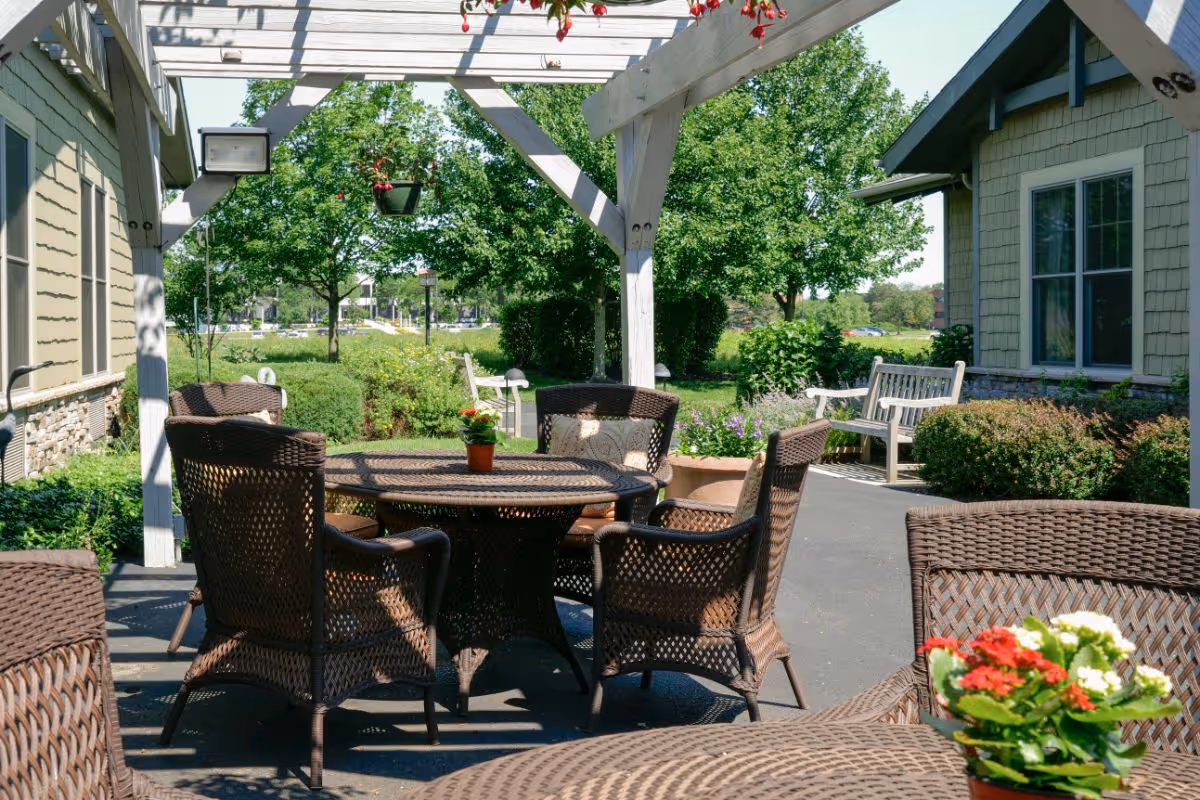 Outdoor patio with wicker tables and chairs under a pergola, surrounded by landscaping and nearby buildings.