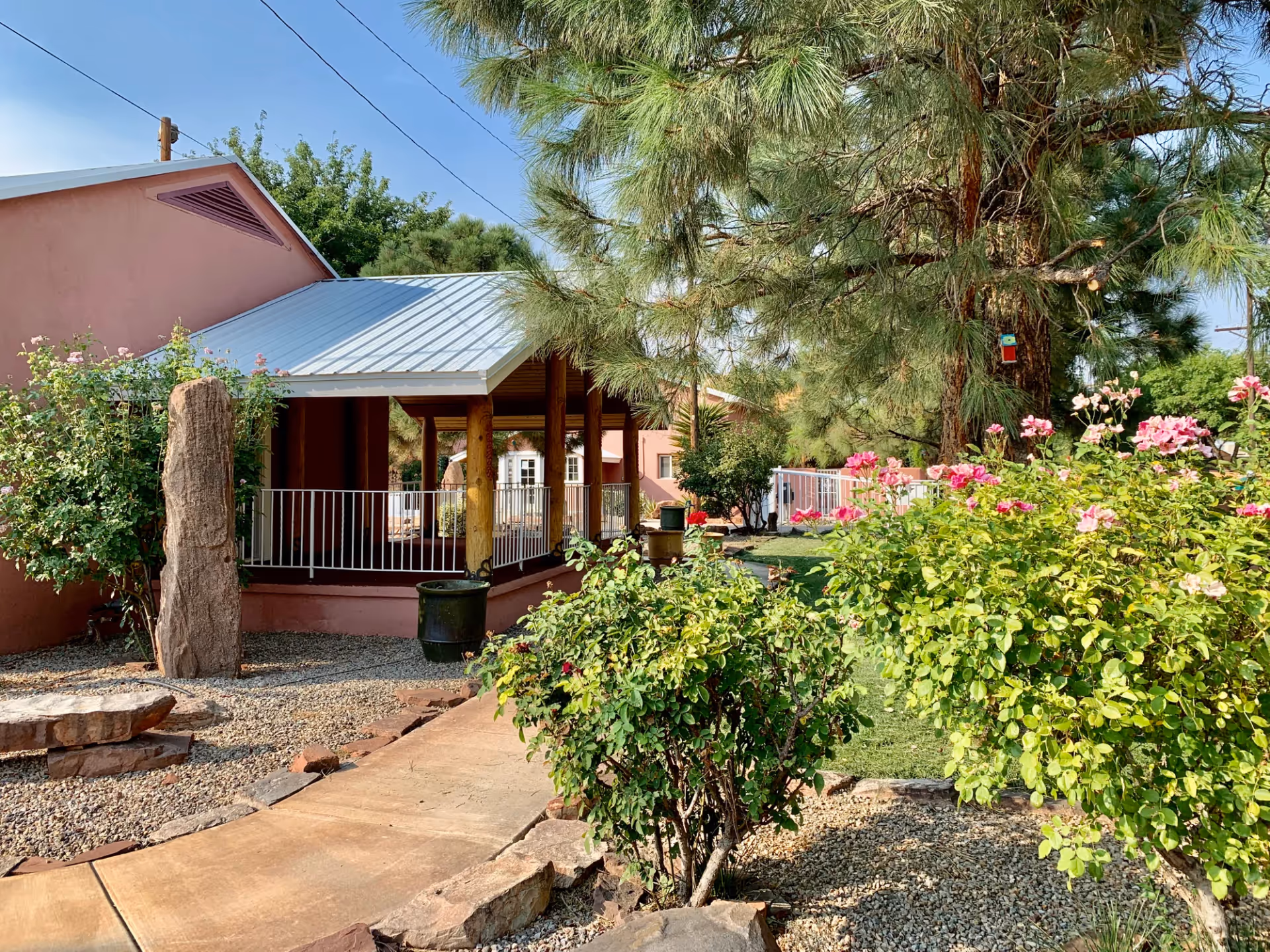 Outdoor garden area at Casa de Rosa Assisted Living with a paved walkway, green bushes with pink flowers, a large tree, and a covered patio attached to a pink building under a clear blue sky.