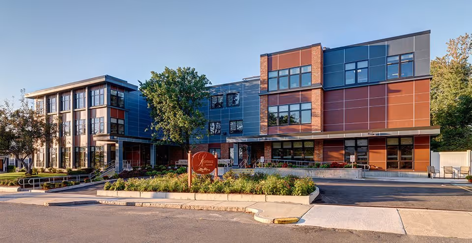 Exterior view of a modern three-story building with large windows and a mix of brick and metal paneling. There is a landscaped area with greenery and a sign in front of the building. The sky is clear and the scene is well-lit by natural daylight.