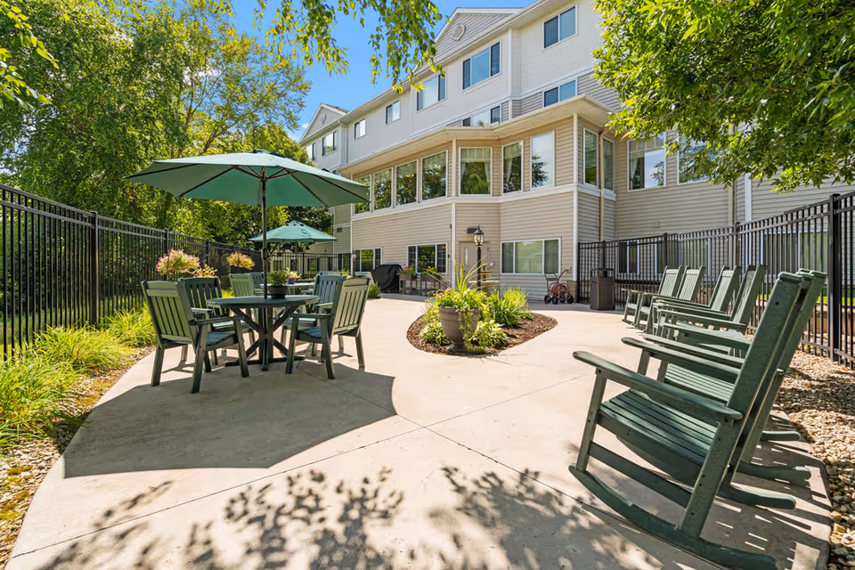 Outdoor patio area at Cornerstone Assisted Living & Memory Care featuring green Adirondack chairs and tables with green umbrellas. The patio is surrounded by a black metal fence and greenery, with a multi-story beige building in the background under a clear blue sky.