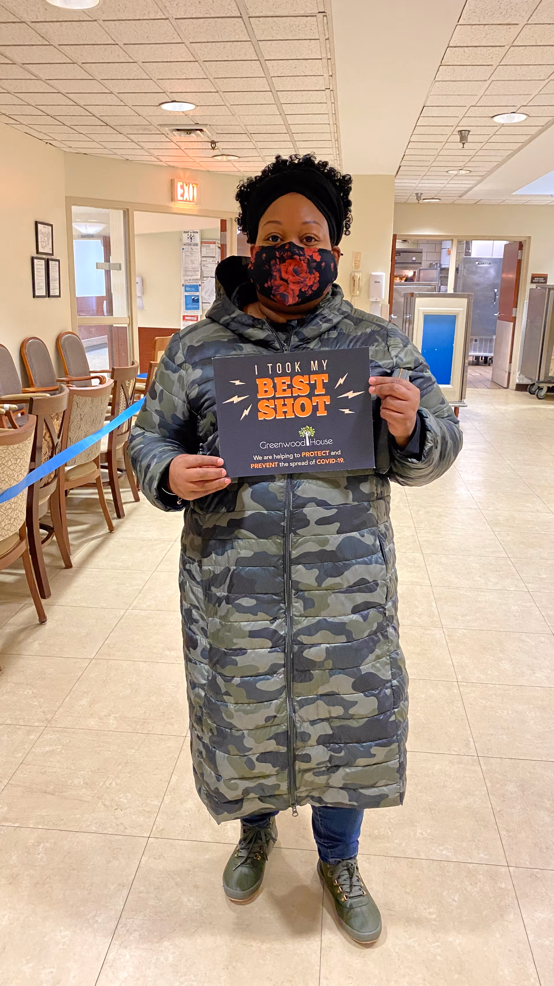A person wearing a camouflage-patterned long coat, green shoes, and a floral face mask stands indoors holding a sign that reads 'I TOOK MY BEST SHOT. Greenwood House. We are helping to PROTECT and PREVENT the spread of COVID-19.' The background shows chairs lined up against the wall and an exit sign above a door.