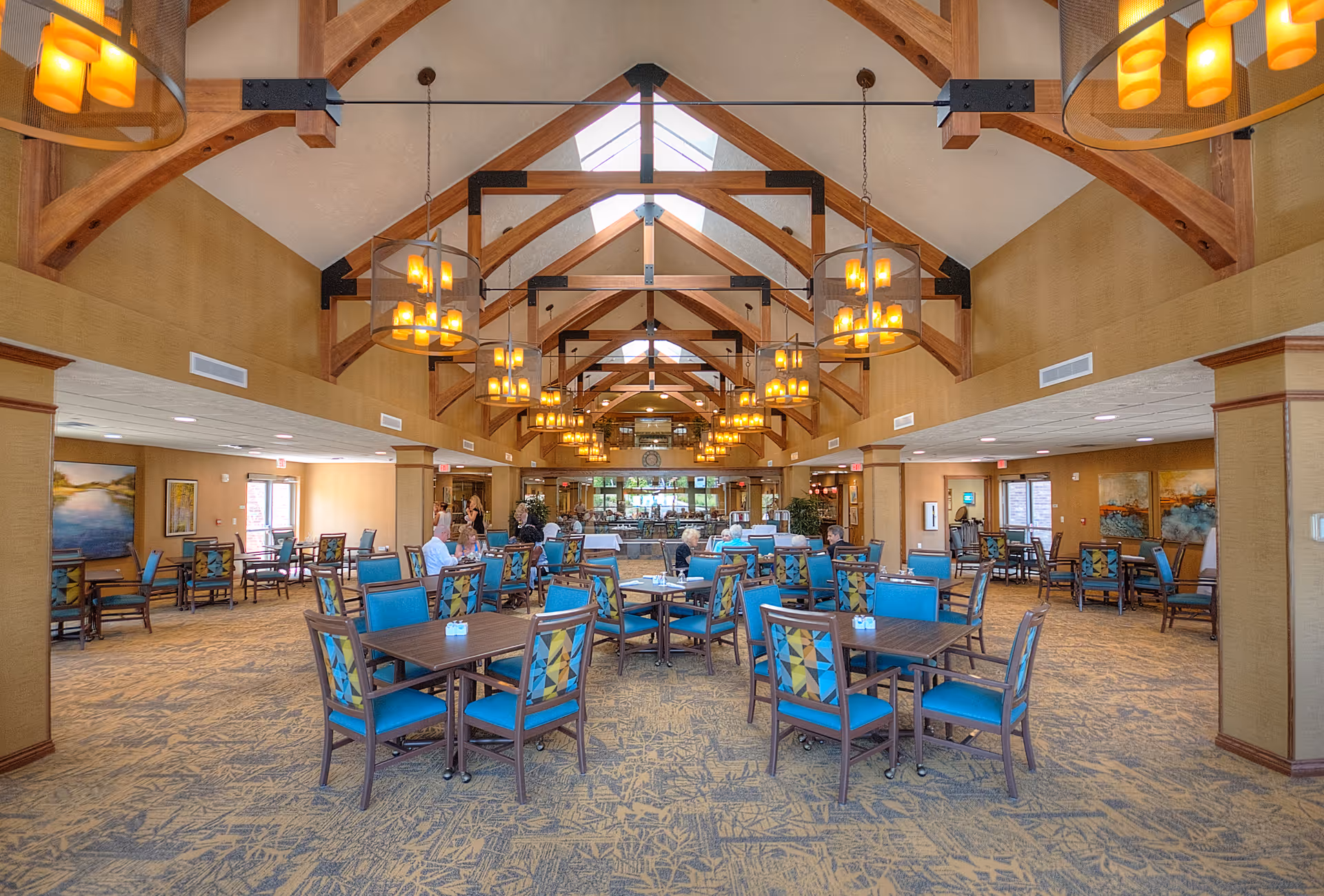 Spacious dining room with multiple tables and blue-upholstered chairs beneath vaulted wooden beams and hanging chandeliers.