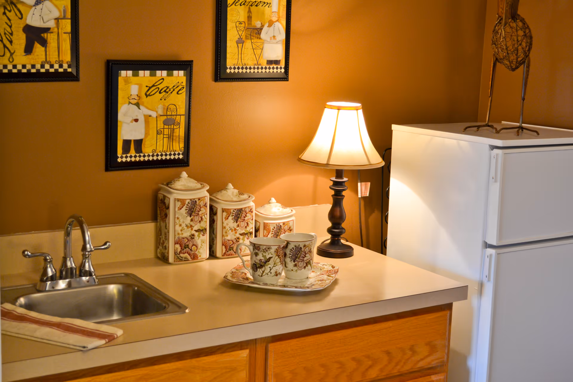 Kitchenette counter with a sink, floral canisters, a teacup on a tray, a lamp, and a white refrigerator against a warm brown wall.