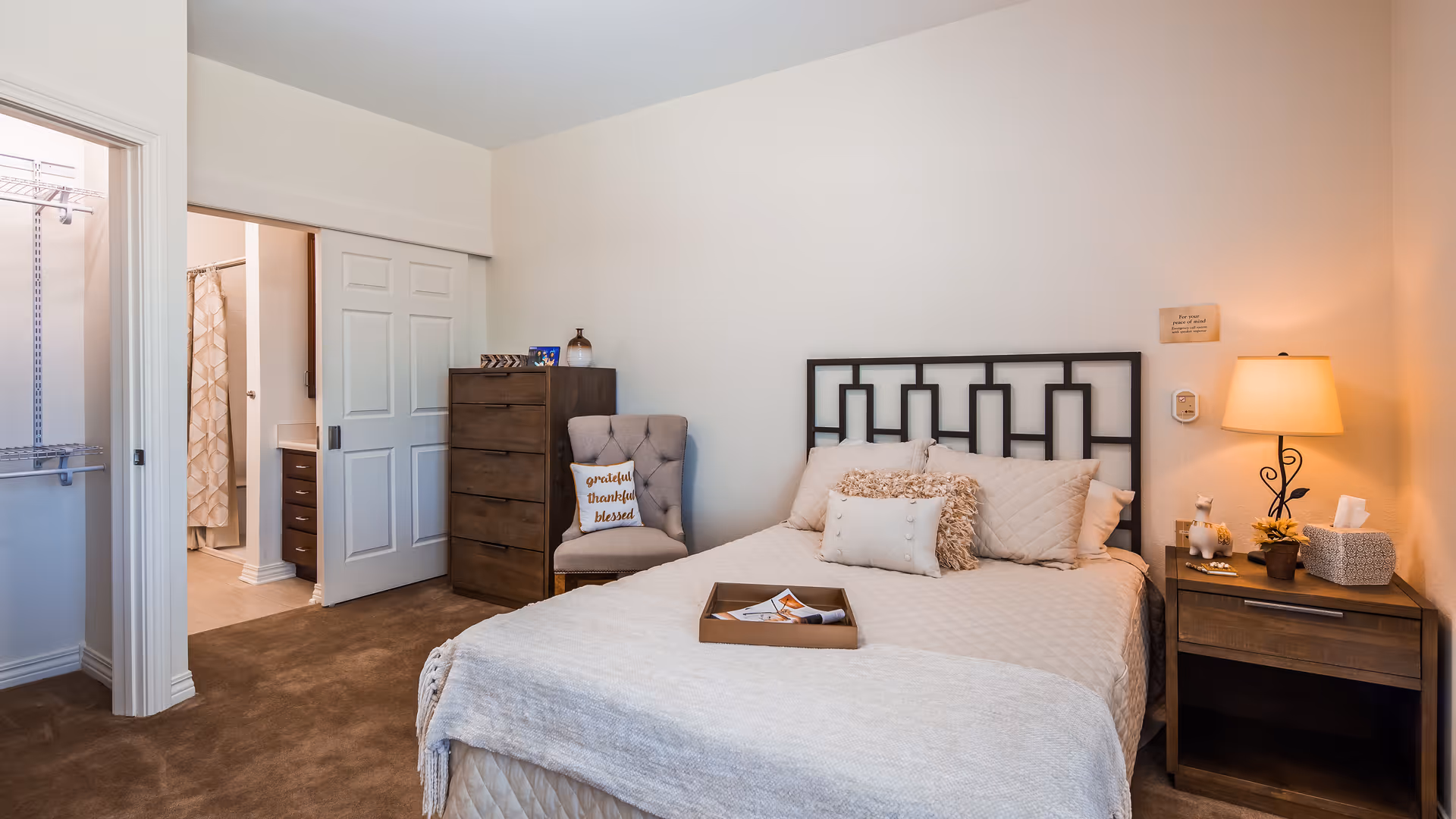 A cozy bedroom with a neatly made bed featuring multiple pillows and a tray on top. To the right of the bed is a wooden nightstand with a lamp, tissue box, and small decorative items. A cushioned chair with a pillow that reads 'grateful thankful blessed' is placed next to a tall wooden dresser. The room has beige carpet and light-colored walls. An open doorway reveals a walk-in closet and a bathroom with a shower curtain.