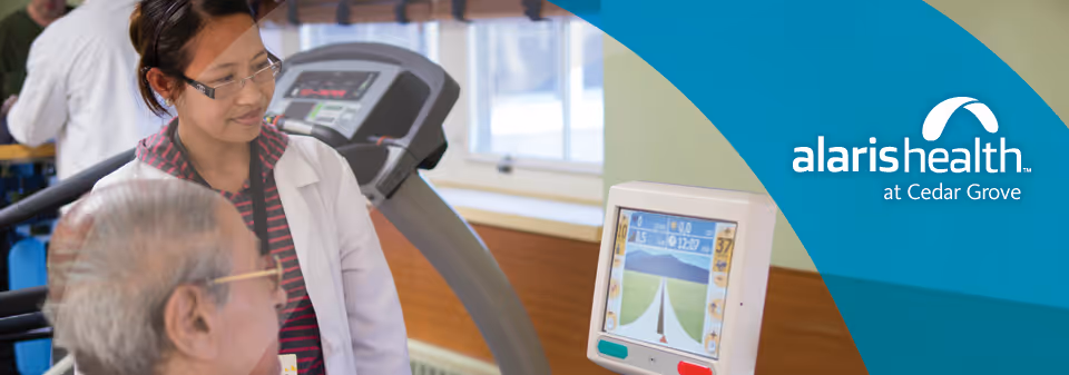 A healthcare professional in a white coat and glasses is assisting an elderly man who is using a treadmill with a digital screen displaying a virtual road and landscape. The setting appears to be a rehabilitation or fitness area within a healthcare facility.