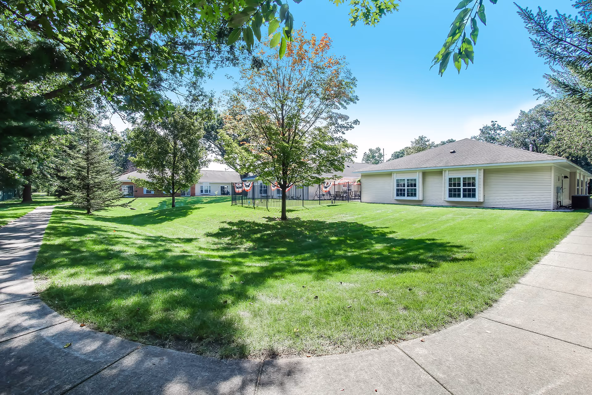 A bright outdoor scene at Ludington Woods Assisted Living and Memory Care showing a well-maintained grassy lawn with several trees casting shadows. There is a paved walkway curving through the green space, and single-story buildings with white siding and multiple windows are visible in the background under a clear blue sky.