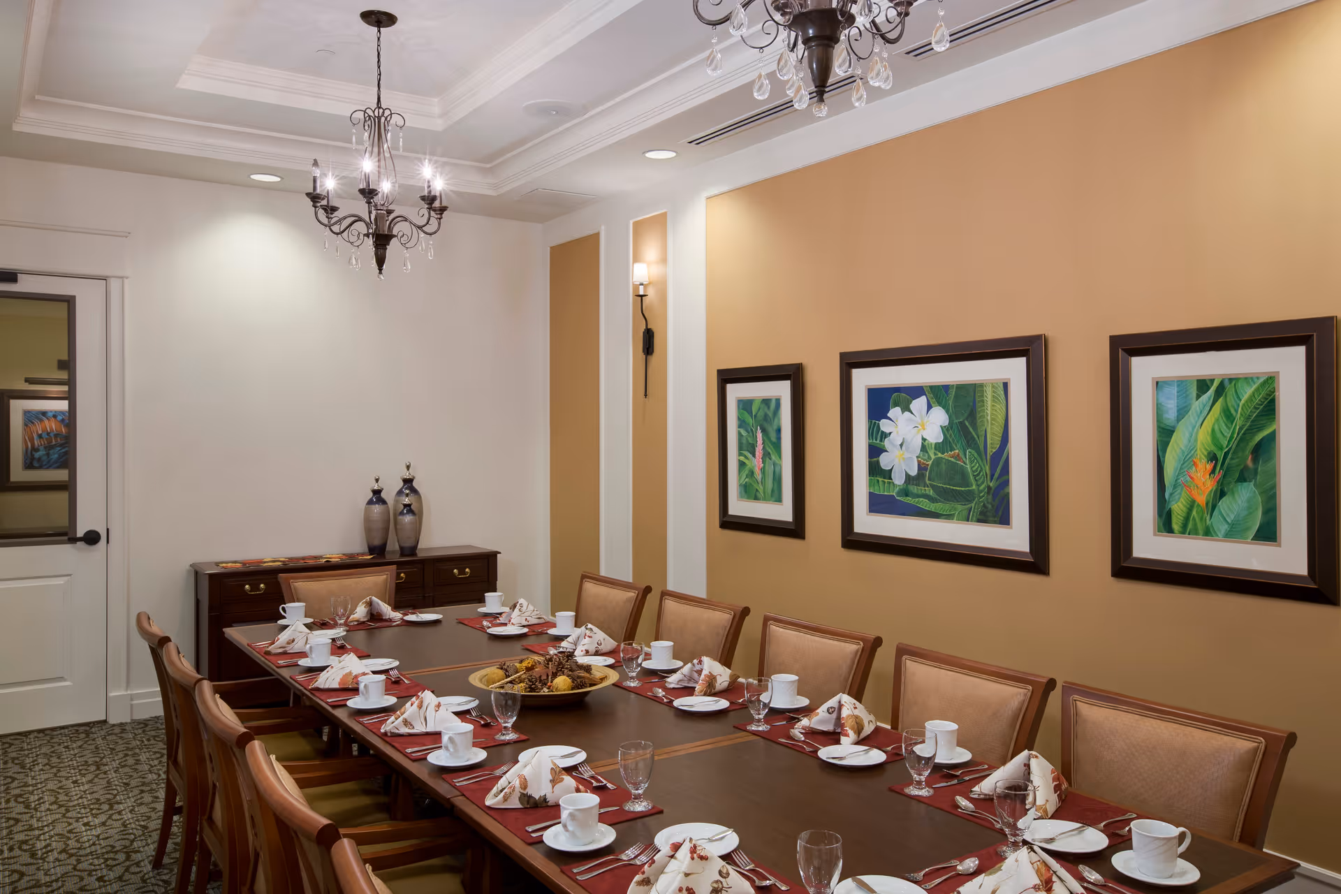 A formal dining room with a long wooden table set for a meal. The table has white plates, cups, glasses, and folded napkins on red placemats. There are twelve wooden chairs with cushioned seats around the table. The walls are painted beige and white, decorated with three framed botanical paintings. A chandelier hangs from the ceiling, and a sideboard with decorative vases is against one wall.
