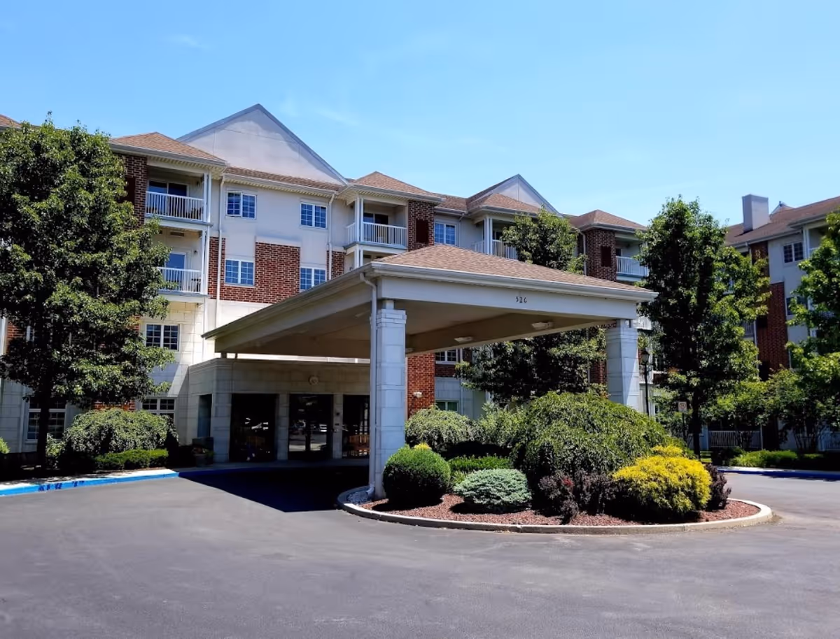 Exterior view of Moravian Village of Bethlehem, showing a multi-story residential building with balconies, a covered entrance with pillars, and landscaped greenery including trees and shrubs under a clear blue sky.