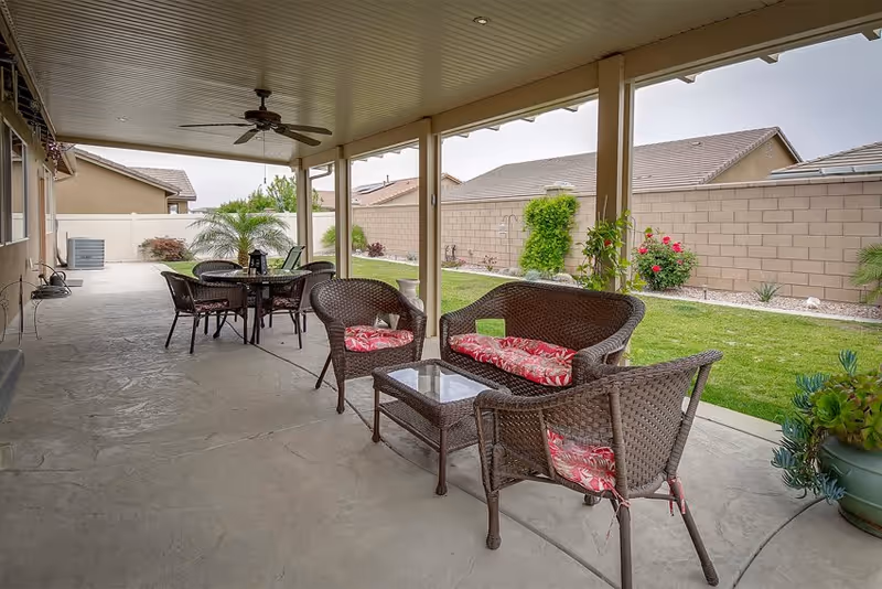 Covered backyard patio with wicker seating and cushions, a dining table, ceiling fan, and a view of a grassy yard and block wall.
