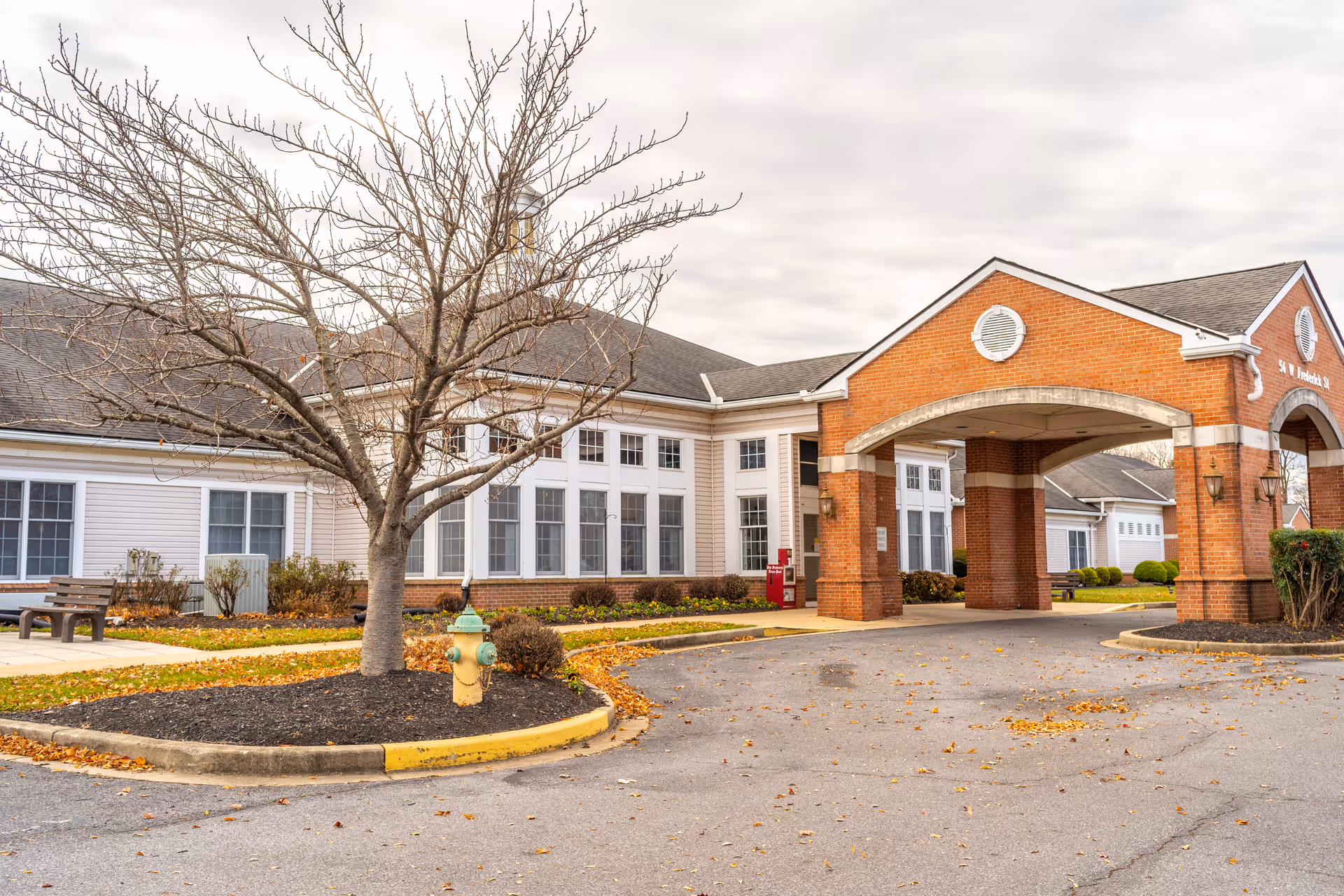Exterior view of Autumn Lake Healthcare at Glade Valley showing a brick entrance archway with a covered driveway, a leafless tree in a mulched island with a fire hydrant, and a building with multiple windows and white siding under a cloudy sky.
