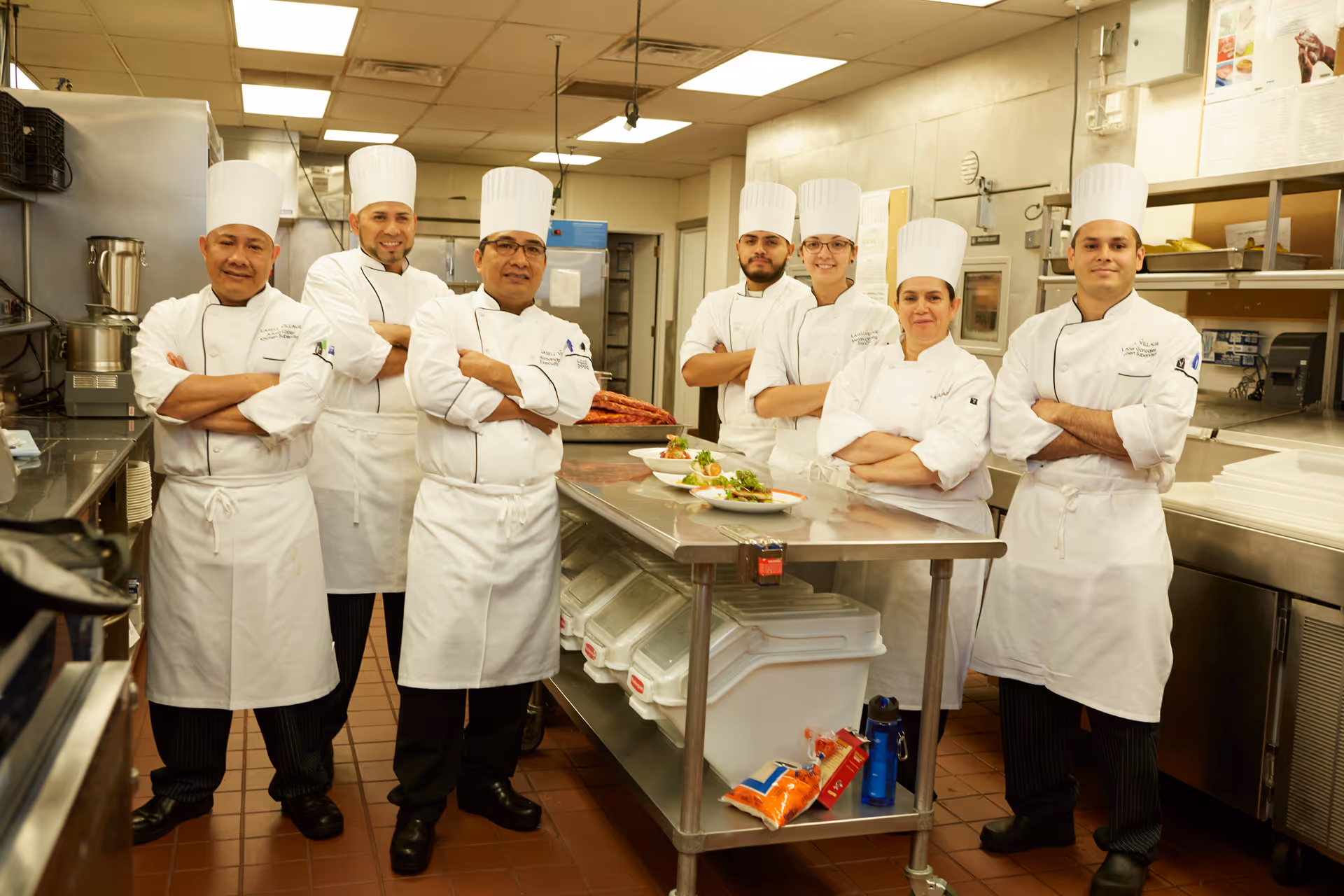 Seven chefs wearing white uniforms and tall white chef hats stand in a commercial kitchen with arms crossed, posing confidently behind a stainless steel table with plated dishes.