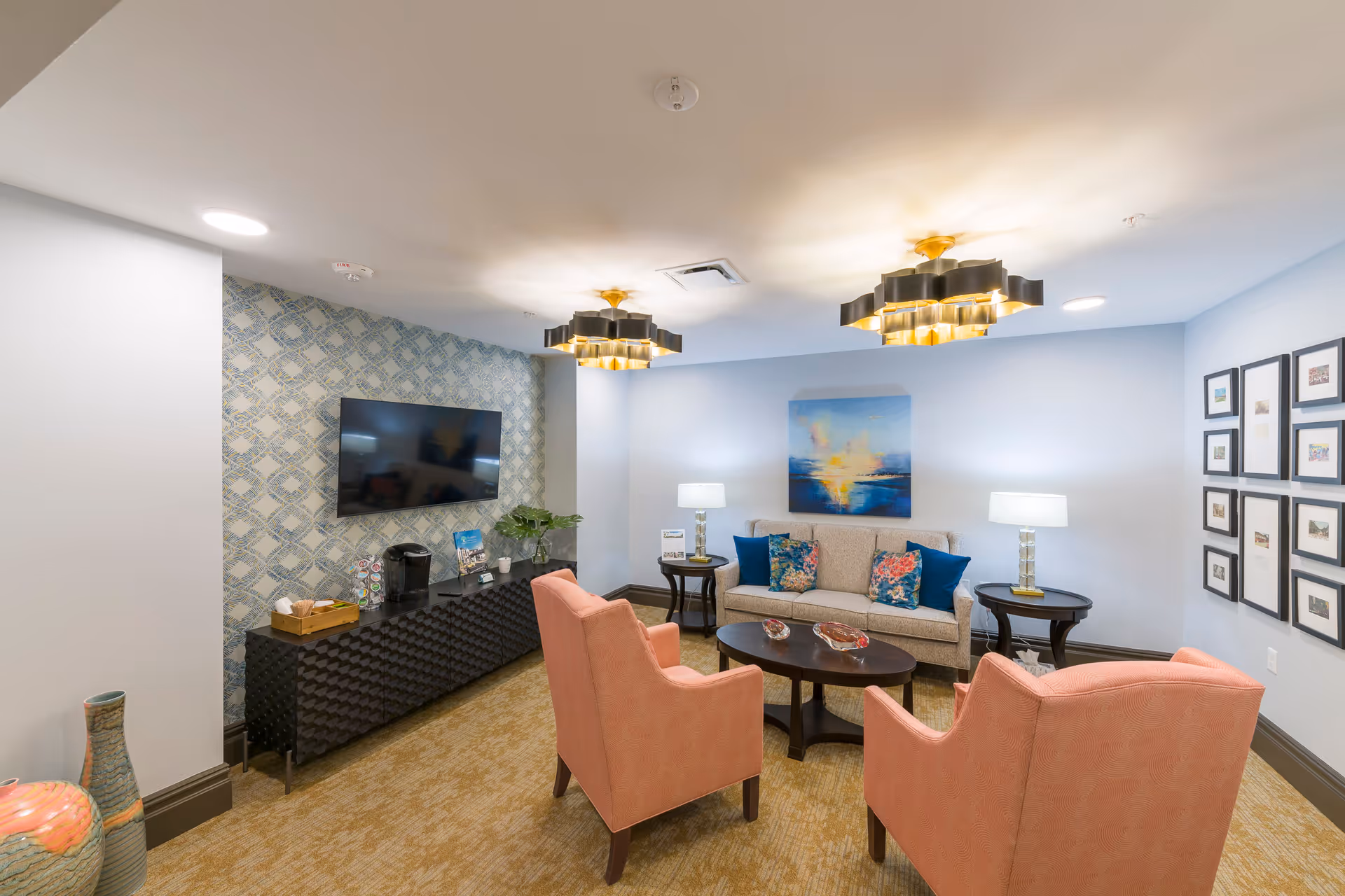 Well-lit communal living room with a sofa, two coral armchairs around a coffee table, wall-mounted TV and decorative artwork.