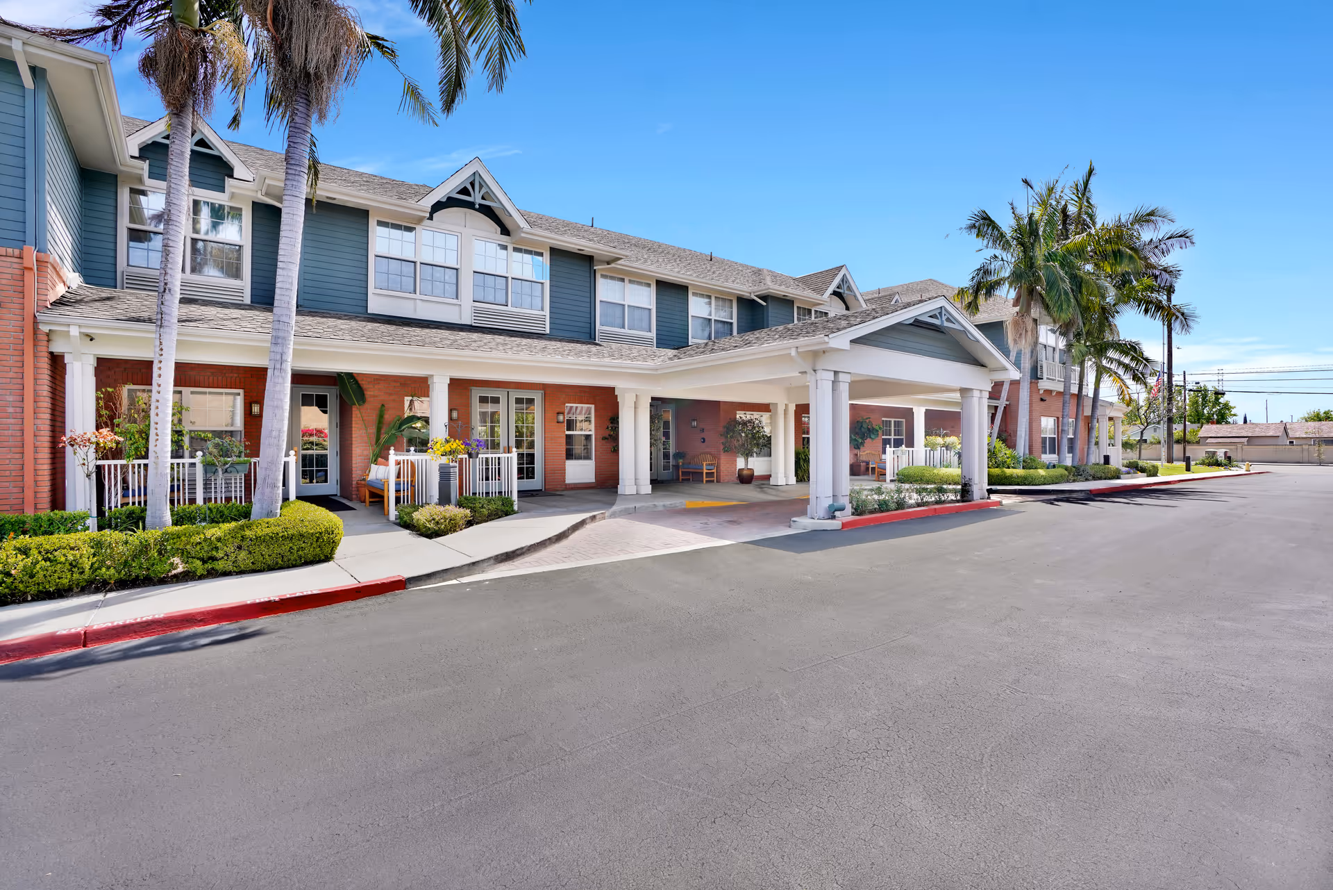 Exterior view of Kirkwood Orange senior living facility showing a two-story building with a covered entrance, palm trees, and well-maintained landscaping under a clear blue sky.