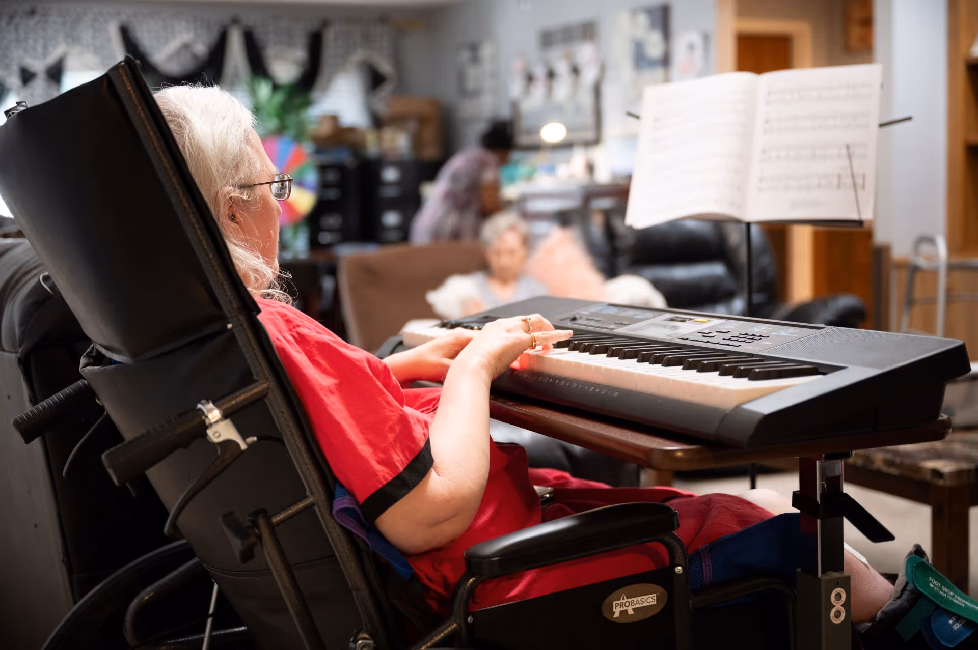 An elderly woman in a wheelchair playing an electronic keyboard with sheet music in front of her, in a cozy living room setting with another person blurred in the background.