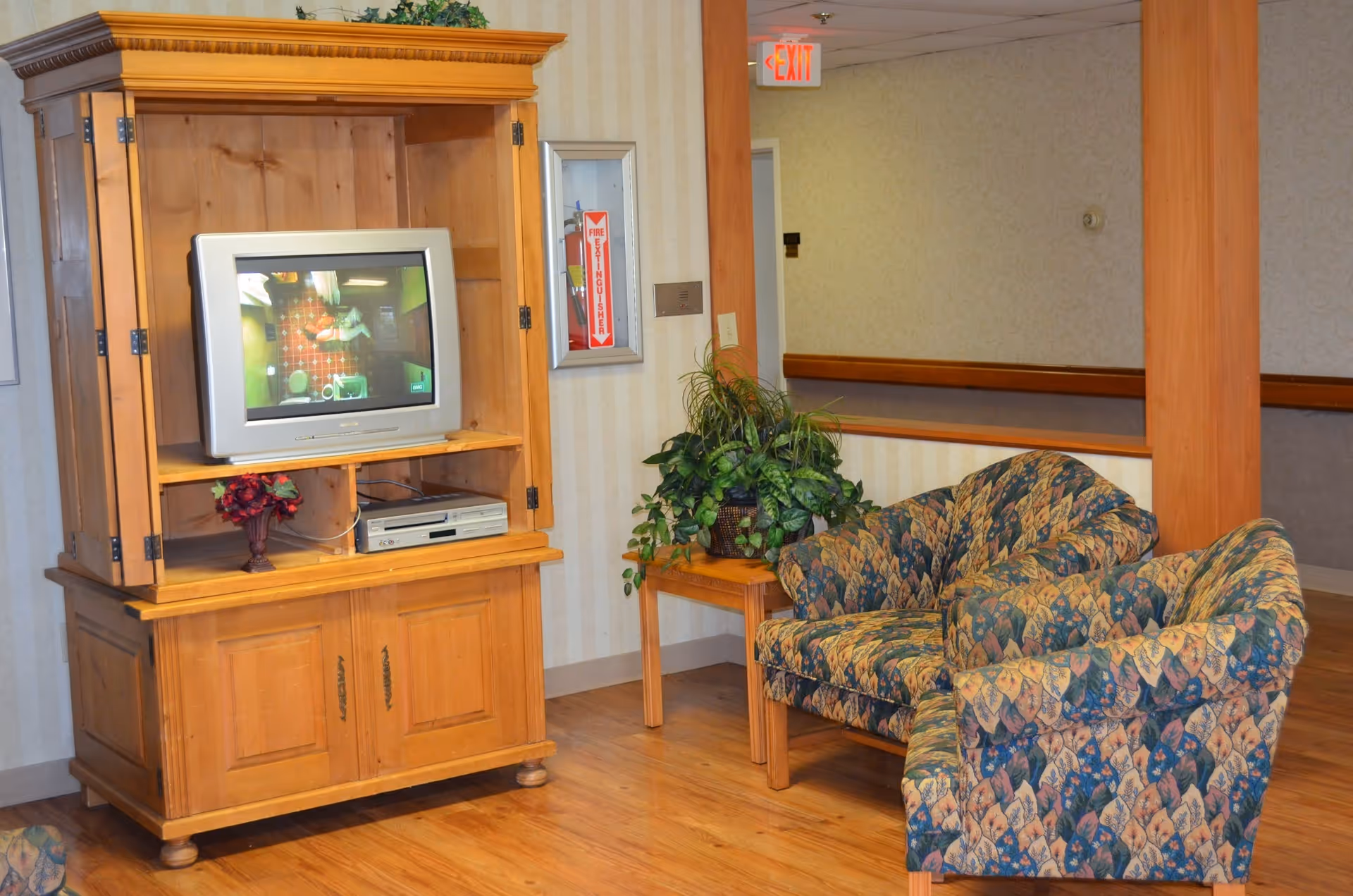 A cozy lounge area with a wooden TV cabinet holding a CRT television, patterned loveseat chairs, a side table with a potted plant, and hardwood floors.