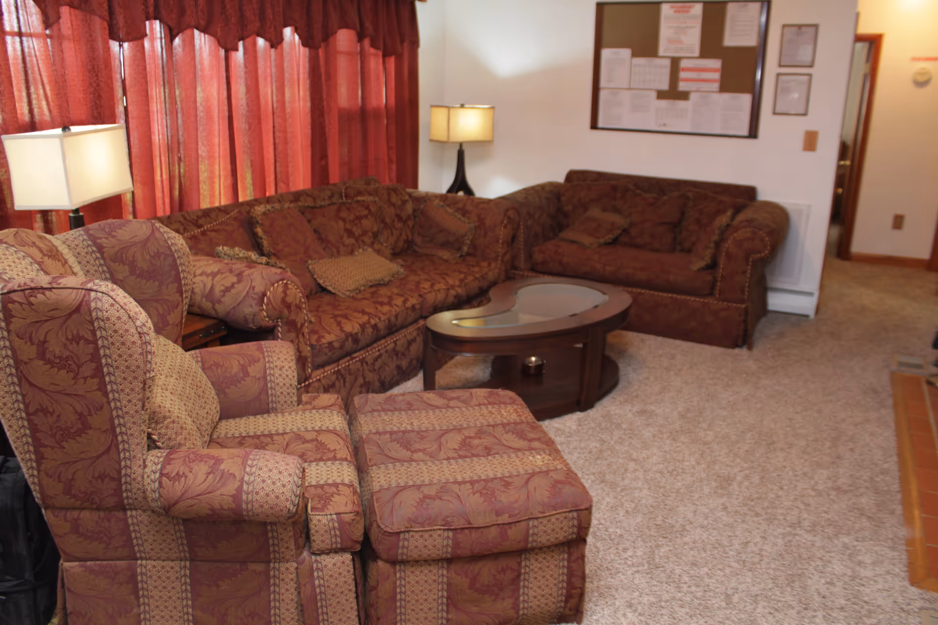 Cozy living room with patterned armchairs and ottoman, a sectional sofa, coffee table, lamps, and red curtains.