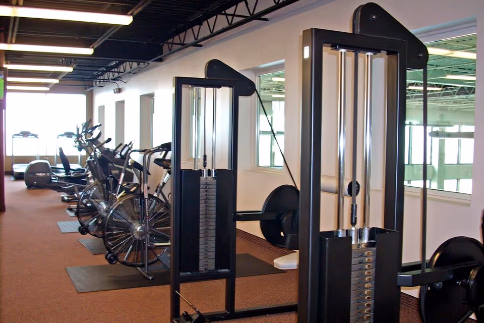 Indoor fitness room with exercise equipment including stationary bikes, treadmills, and weight machines, with large windows allowing natural light.