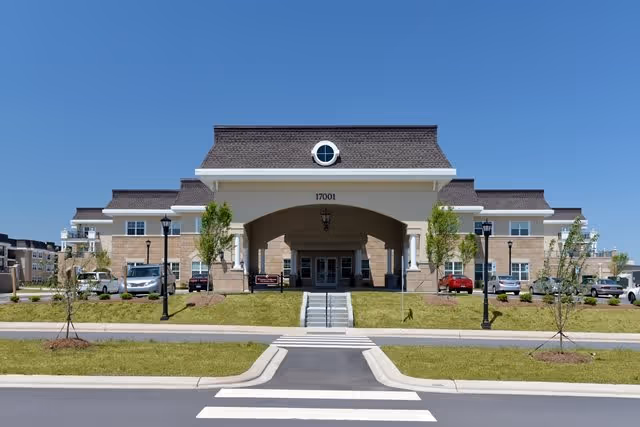Front exterior view of the SearStone facility building with a large covered entrance, multiple windows, a circular window above the entrance, and a clear blue sky. There are small trees and grass in front, along with a crosswalk leading to the entrance.