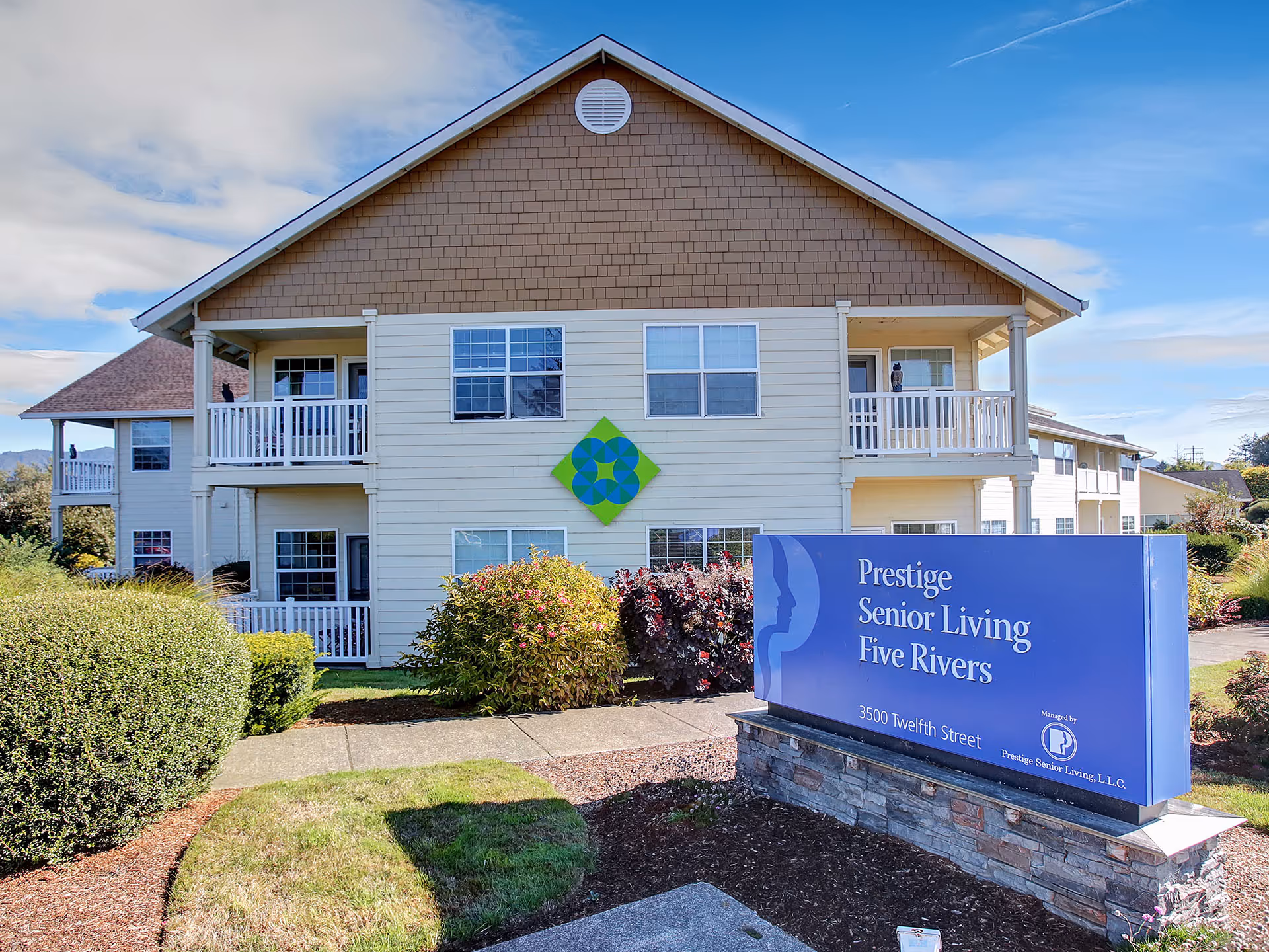 Exterior view of a two-story senior living facility building with beige siding and brown shingles. The building has balconies with white railings and several windows. In front of the building, there is a blue sign that reads 'Prestige Senior Living Five Rivers' along with the address '3500 Twelfth Street' and a logo. The area around the building is landscaped with bushes, grass, and a clear blue sky overhead.