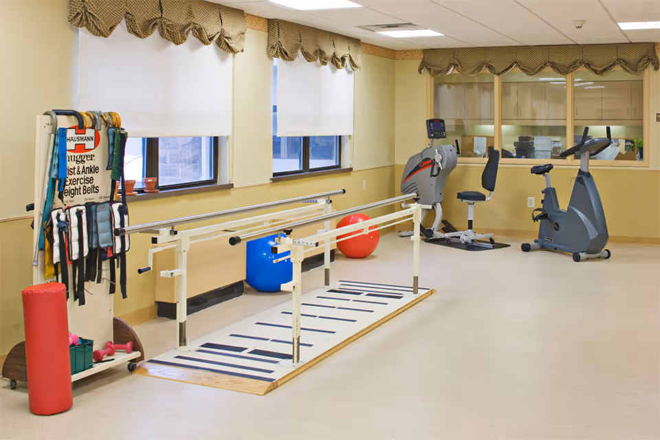 A rehabilitation or exercise room with parallel bars for walking practice, exercise balls, stationary exercise bikes, and various exercise belts and weights hanging on a rack. The room has beige walls, windows with white roller blinds, and a large window looking into another room.