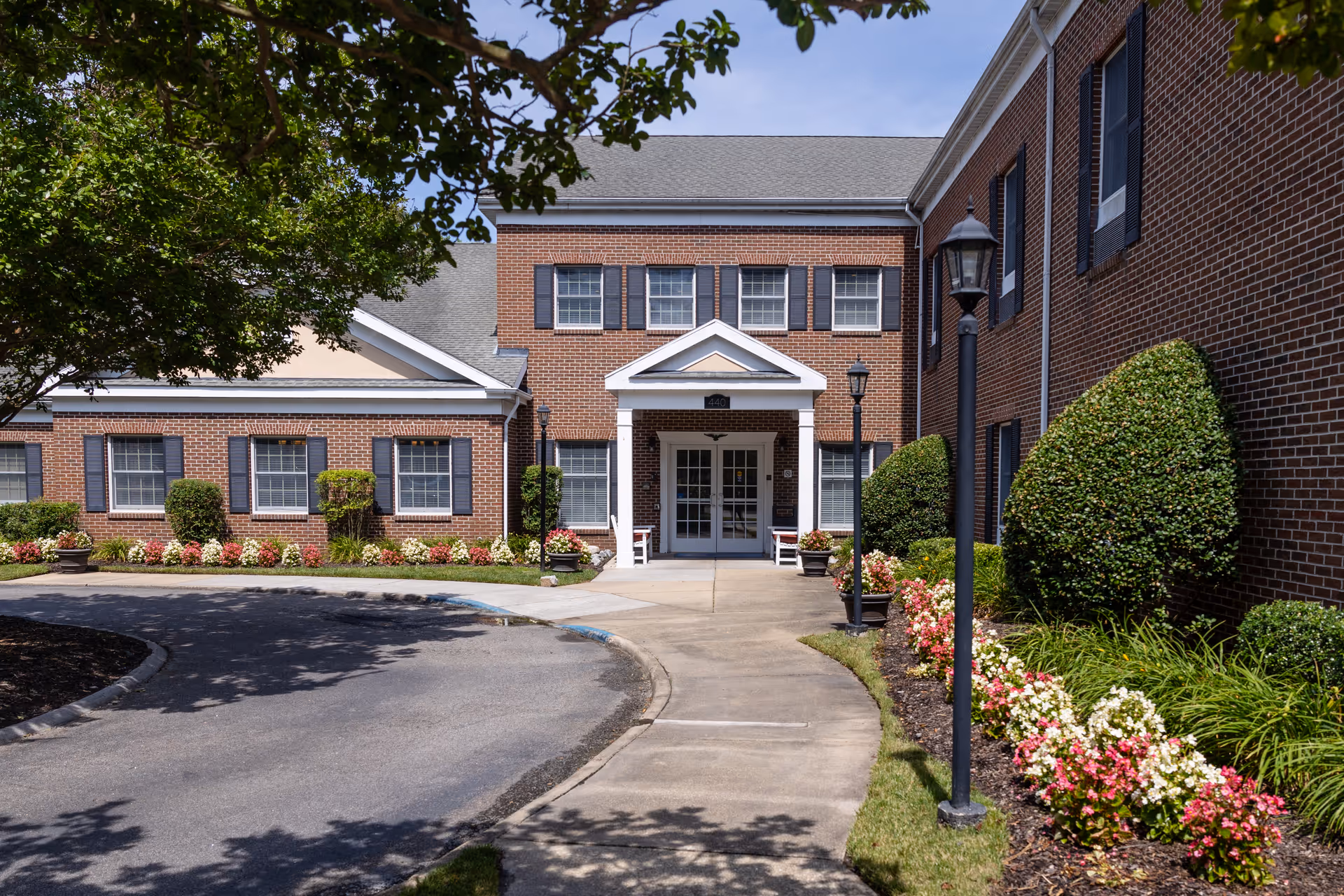 Front exterior view of a brick senior living facility building with a paved driveway and a sidewalk leading to double glass doors under a white portico. The building has multiple windows with black shutters, well-maintained landscaping with trimmed bushes and colorful flowers, and black lamp posts along the sidewalk.