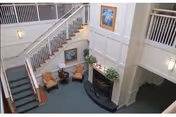 Interior view of a senior living facility common area with a staircase, two armchairs, a small round table, a fireplace with plants on the mantel, and framed artwork on the walls. The space has a high ceiling and railings on the upper level.
