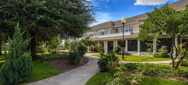 Landscaped courtyard with a curved concrete path, trees and shrubs leading to a two-story building with balconies and a lamppost under a blue sky.