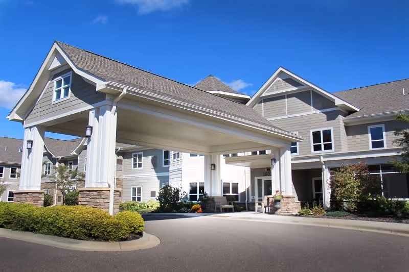 Exterior view of a senior living facility building with a covered entrance driveway, landscaped bushes, and clear blue sky.